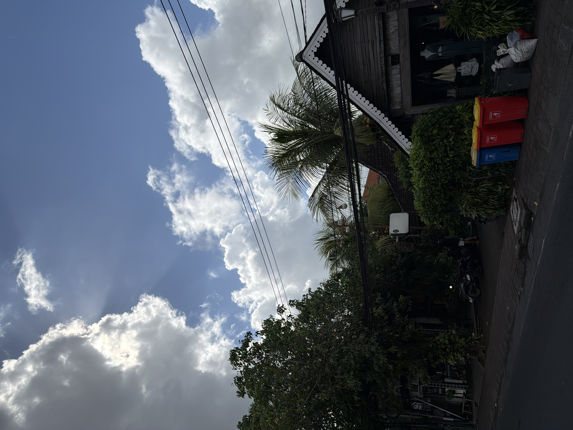 The scene is a street in North Kuta, Indonesia, under a partly cloudy sky. Power lines run diagonally across the frame. A traditional wooden building with a display window stands alongside the street, where trash bins are placed. A palm tree and other dense foliage surround the building. A scooter is parked on the sidewalk. The pavement is dark and possibly wet. No people are clearly visible.