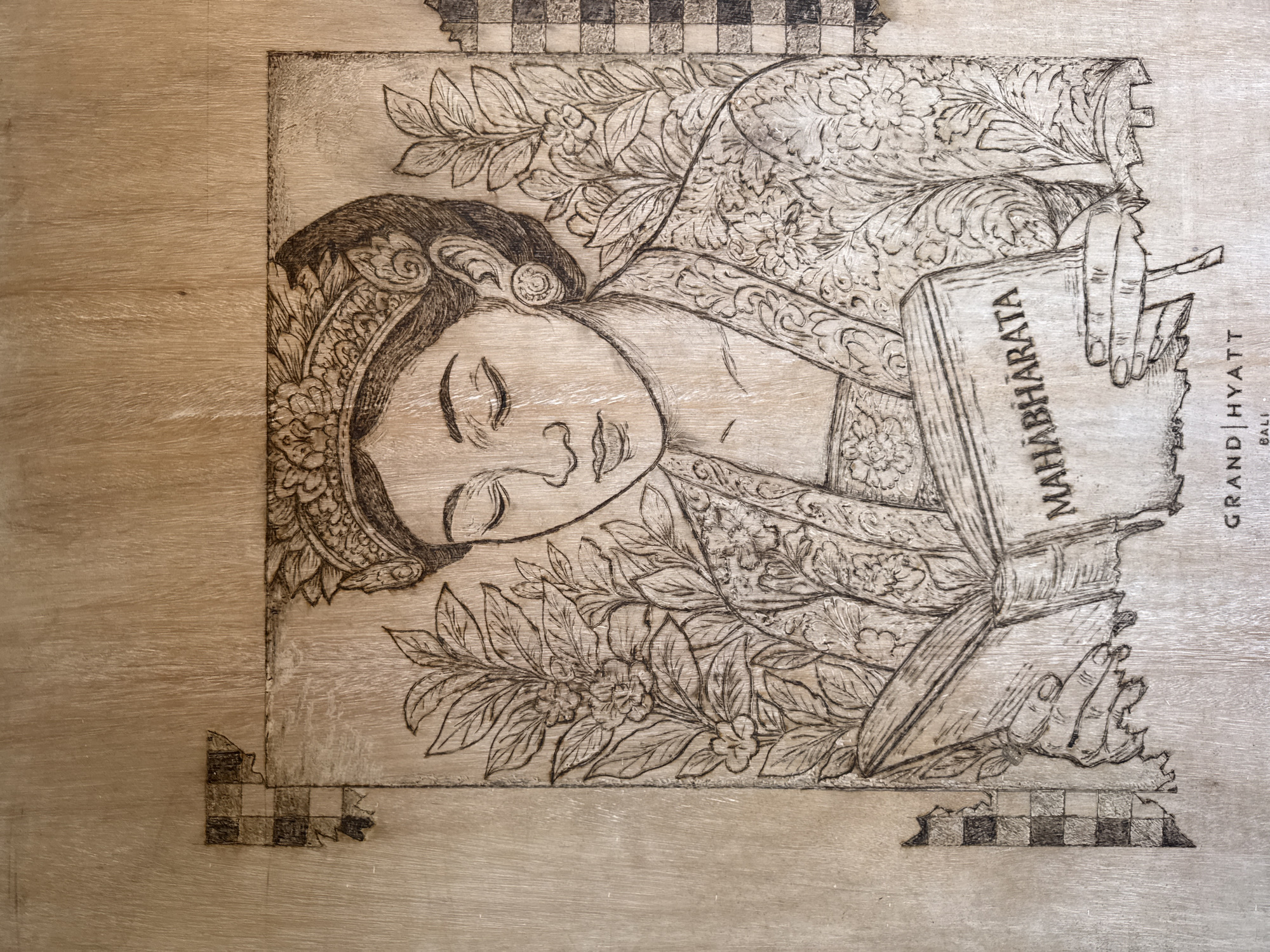 This is an image of a wood carving. The carving depicts a person lying down, holding a book titled "Mahabharata". The carving also includes foliage around the figure. The words "GRAND HYATT" and "BALI" are visible on the right side. The carving is located in Kuta Selatan, Indonesia, as indicated by the text on the carving.