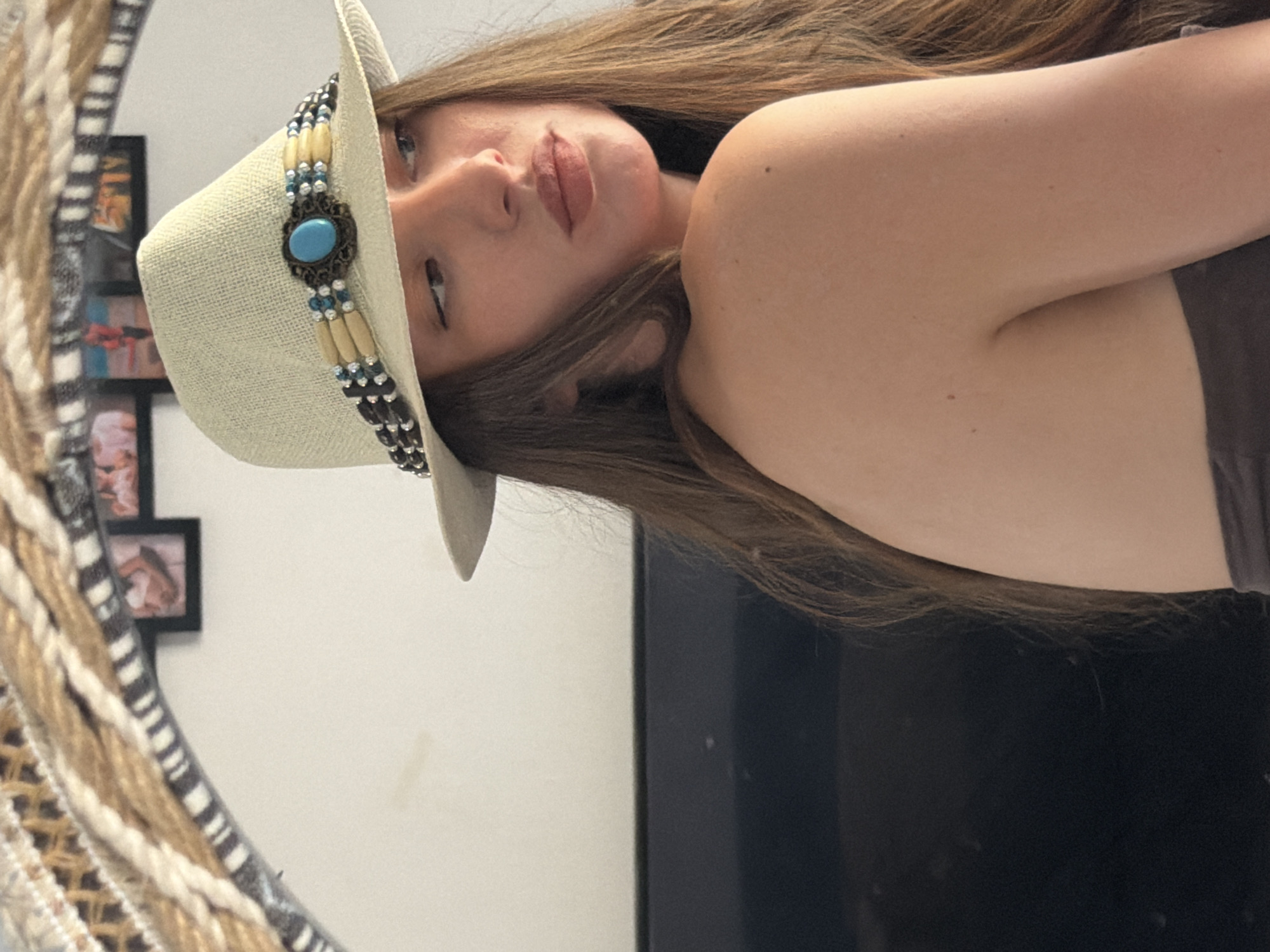 A woman wearing a straw fedora hat with a decorative band is shown from the chest up, facing away from the camera and looking over her shoulder. The hat has a turquoise stone and wooden beads. Her long, brown hair falls over her shoulders. She is wearing a dark-colored strapless top. The background consists of a white wall with framed pictures and a decorative woven tapestry above. A dark, flat surface, possibly a television, is visible to the left of the woman. The setting appears to be an indoor environment.