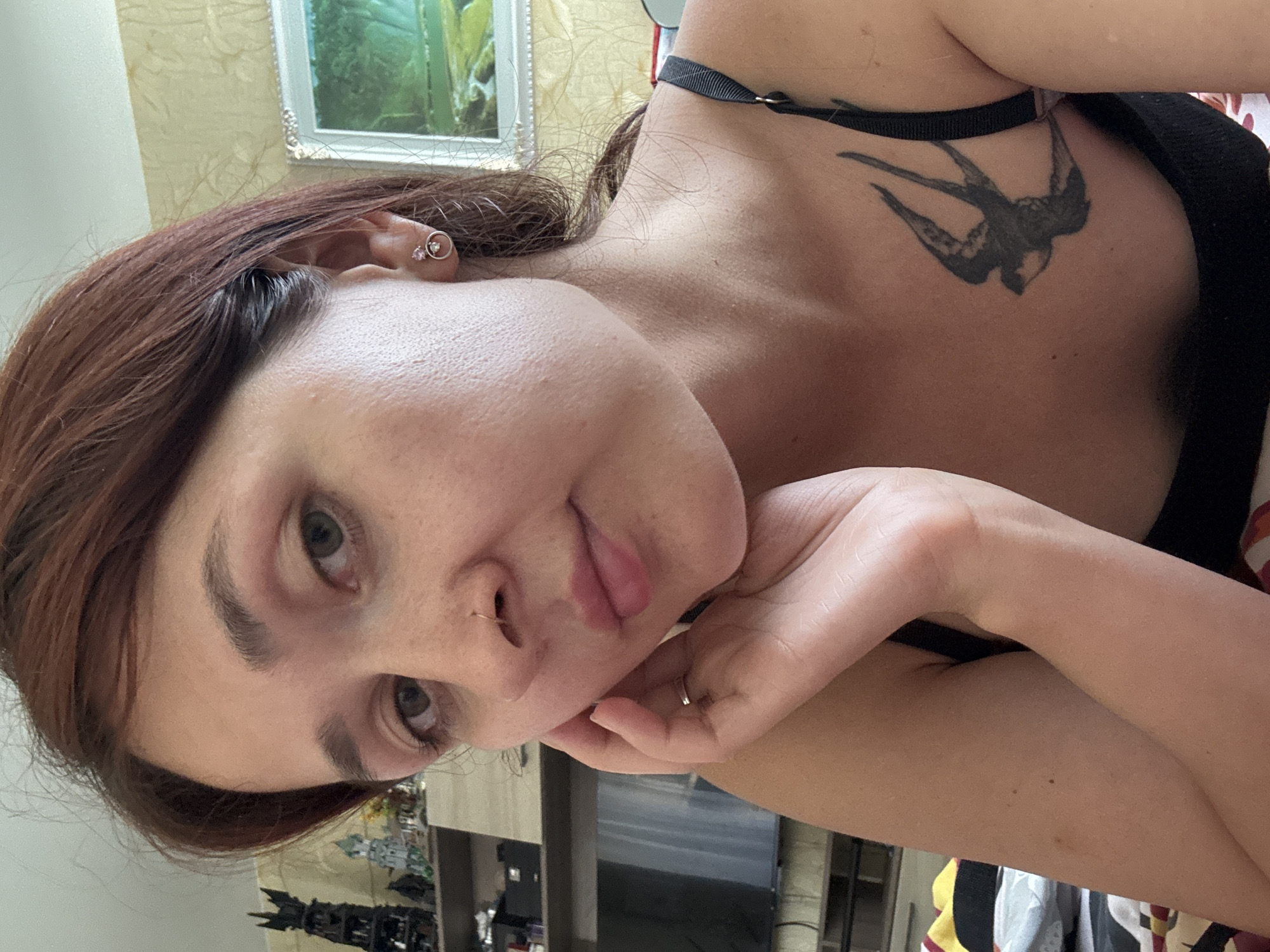 A woman with auburn hair and light skin is shown in a close-up portrait, resting her head on her hand. She is wearing a black bra, and a tattoo of a swallow is visible on her chest. She has a nose ring and small earrings. The background includes a painting on the wall and shelves with various objects. The lighting suggests an indoor setting, likely a residential room. There are no clear indicators of the specific location within Naberezhnye Chelny, Russia, or any city.