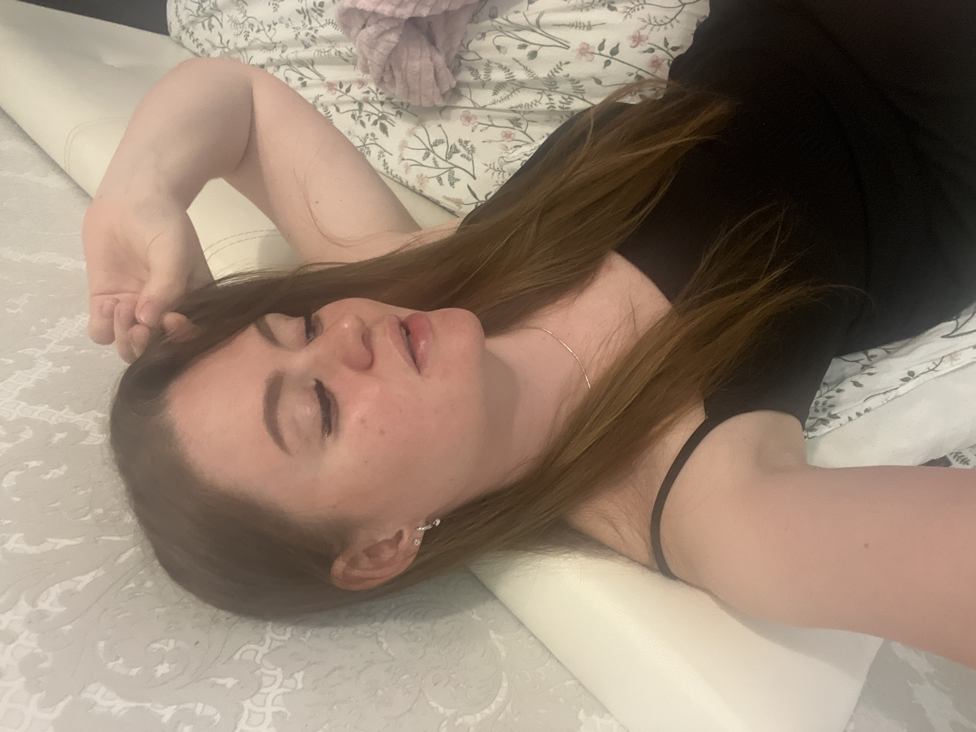 A woman is lying on a patterned white bedspread. She has long, brown hair and is wearing a black top. Her eyes are closed, and she is lying on her side with one arm above her head. A white, cylindrical pillow supports her head and neck. The location is unknown.