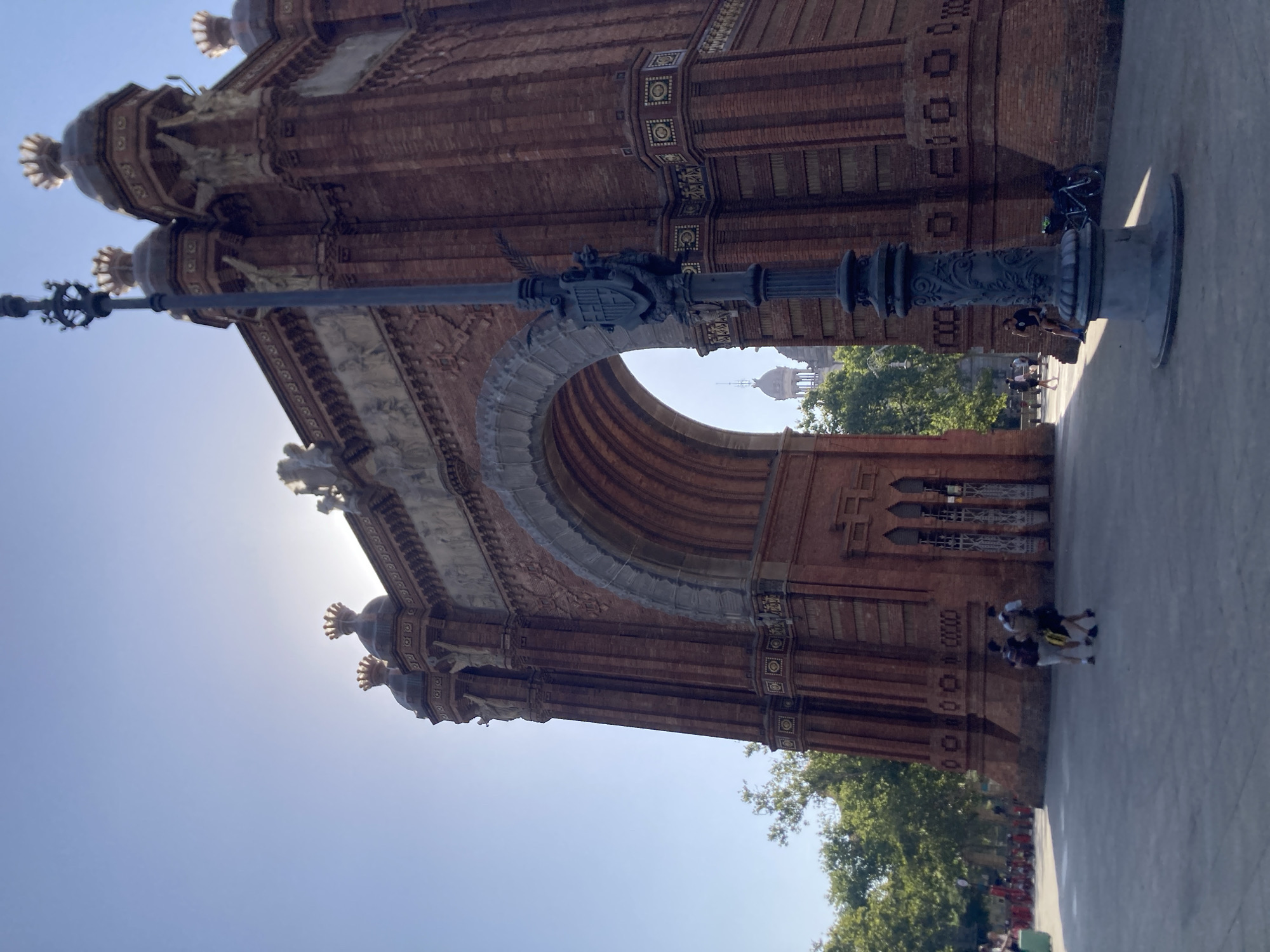 This media file captures a sunny day at the Arc de Triomf in Barcelona, Spain. The iconic brick archway dominates the frame, showcasing its intricate architectural details, including decorative friezes and ornate finials. A lamppost with a decorative top stands in the foreground, casting a shadow on the paved ground.
Several people are visible in the scene. Some are walking and conversing, while others appear to be taking a break. A bicycle is parked near the base of the arch. In the background, lush green trees and the clear blue sky indicate pleasant weather. The sun is high in the sky, suggesting it is likely midday. The overall atmosphere appears relaxed and leisurely, typical of a tourist attraction on a bright day. The image is oriented vertically, with the sky occupying the left side of the frame and the monument and ground on the right.