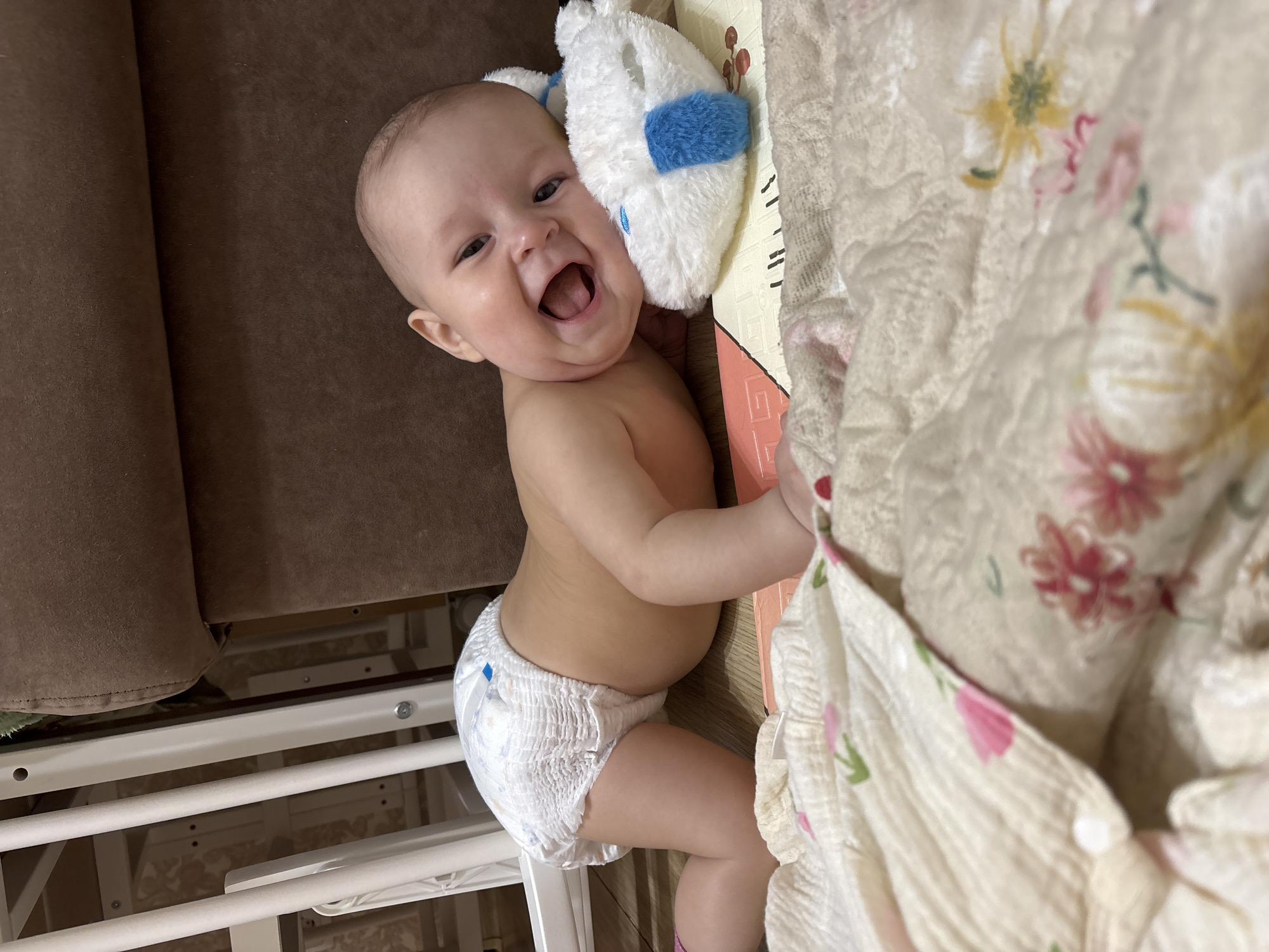 A baby is lying on a surface with a floral-patterned cloth nearby, appearing to be a blanket. The baby is wearing a diaper and is smiling broadly. A white plush toy with blue accents is next to the baby's head. Part of a brown sofa is visible in the background, along with a white metal frame, possibly part of a crib. The location cannot be determined from the image, nor can the city of Sochi, Russia be confirmed.