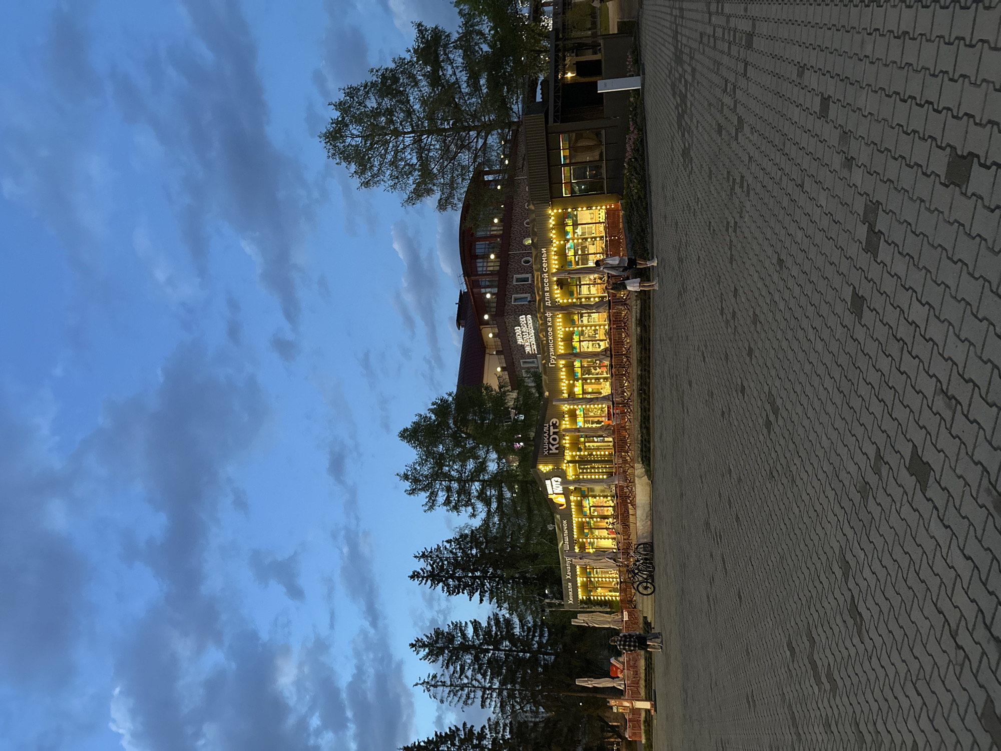 Captured at dusk, the image showcases a street in Yekaterinburg, Russia. A cobblestone walkway extends to the right. A multi-story building borders the path and is brightly lit by strung lights and interior illumination, the storefronts display signs in Russian. Tall evergreen trees and a deciduous tree are interspersed along the storefront. Overhead, the sky is blue, streaked with dark gray clouds. Several bicycles are parked. At least three people are near the building, with two standing by an entrance. Another individual is standing near the tree line.