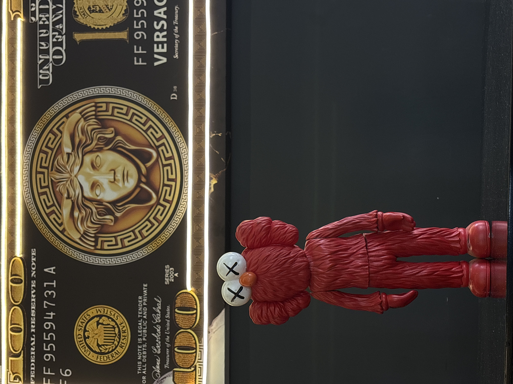 A red KAWS figurine, with "X" marks for eyes, is positioned in front of a framed Versace-themed decorative note. The note is black and gold, mimicking the design of a US dollar bill, but with Versace branding and imagery. It includes details such as "FEDERAL RESERVE NOTE", a series number "FF 95594731 A", and the words "VERSACE" and "Secretary of the Treasury." The environment appears to be indoors, with a dark background and possibly a reflective surface underneath the figurine and framed note. The location is Puebla, Mexico.