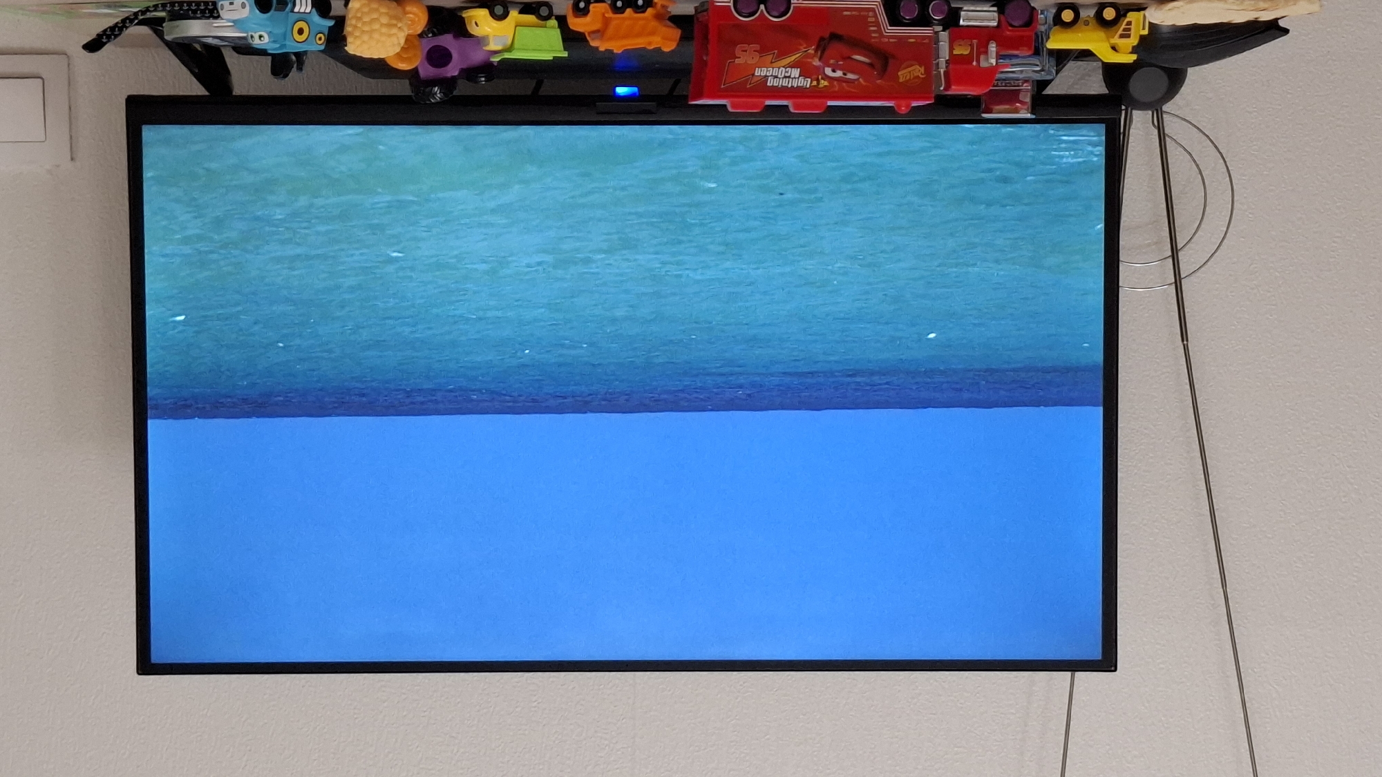 A wall-mounted television displays an image of the sea, appearing to show the surface and underwater view. A shelf above the TV is lined with various colorful toy cars and trucks, including a Lightning McQueen toy. The wall is plain and light-colored. The scene does not provide any information indicating the location is Saint Petersburg, Russia.