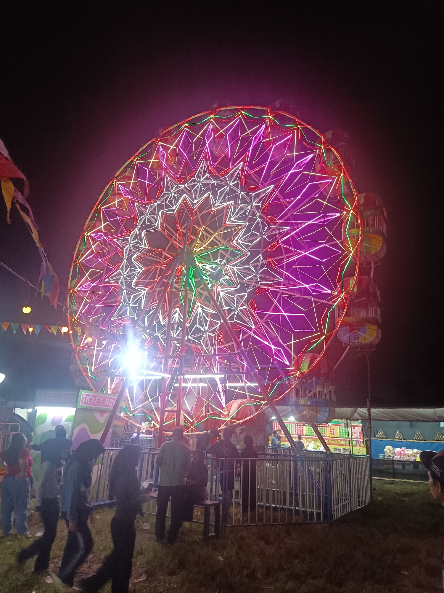 This is a nighttime, low-angle, full shot of a brightly lit Ferris wheel at an outdoor carnival or fair, likely located in Depok, Indonesia. The Ferris wheel is the dominant subject, adorned with intricate patterns of white, pink, and green LED lights. The structure of the wheel is visible, with multiple cars or gondolas attached. In the foreground and midground, several people are present, some walking and others standing in lines or near stalls. A "TICKET BOX" sign is visible on a stall to the left of the Ferris wheel. The ground is covered in grass. Bunting and decorative flags are strung up around the carnival area. The overall atmosphere appears festive and lively.