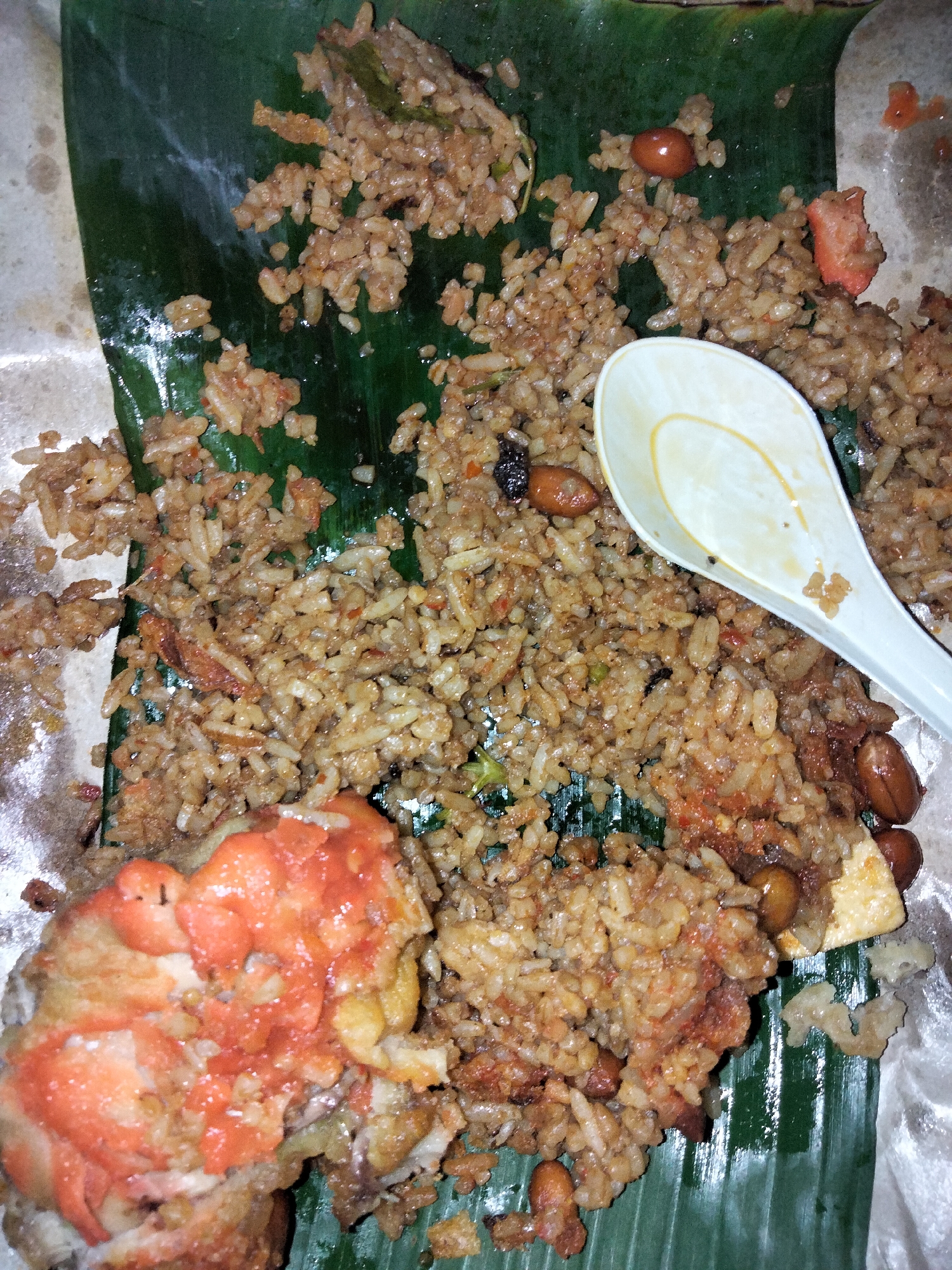 A close-up, overhead view shows a portion of Nasi Uduk, a popular Indonesian rice dish, served on a banana leaf. The rice is a light brown color, mixed with various ingredients such as small pieces of meat, peanuts, and what appears to be chili paste. Some of the rice has spilled onto the surrounding white paper packaging.
In the lower left corner, a piece of fried chicken, coated in a reddish sauce, is visible. A white plastic spoon rests on top of the rice in the upper right quadrant of the image. The banana leaf underneath the food is a vibrant green, with visible veins. The overall impression is of a casually served, hearty meal. The lighting is natural, suggesting it could be daytime. No text is visible in the image. The food is presented in a straightforward manner, hinting at a casual dining experience, possibly from a street vendor or a local eatery in Payagaboh, Indonesia.