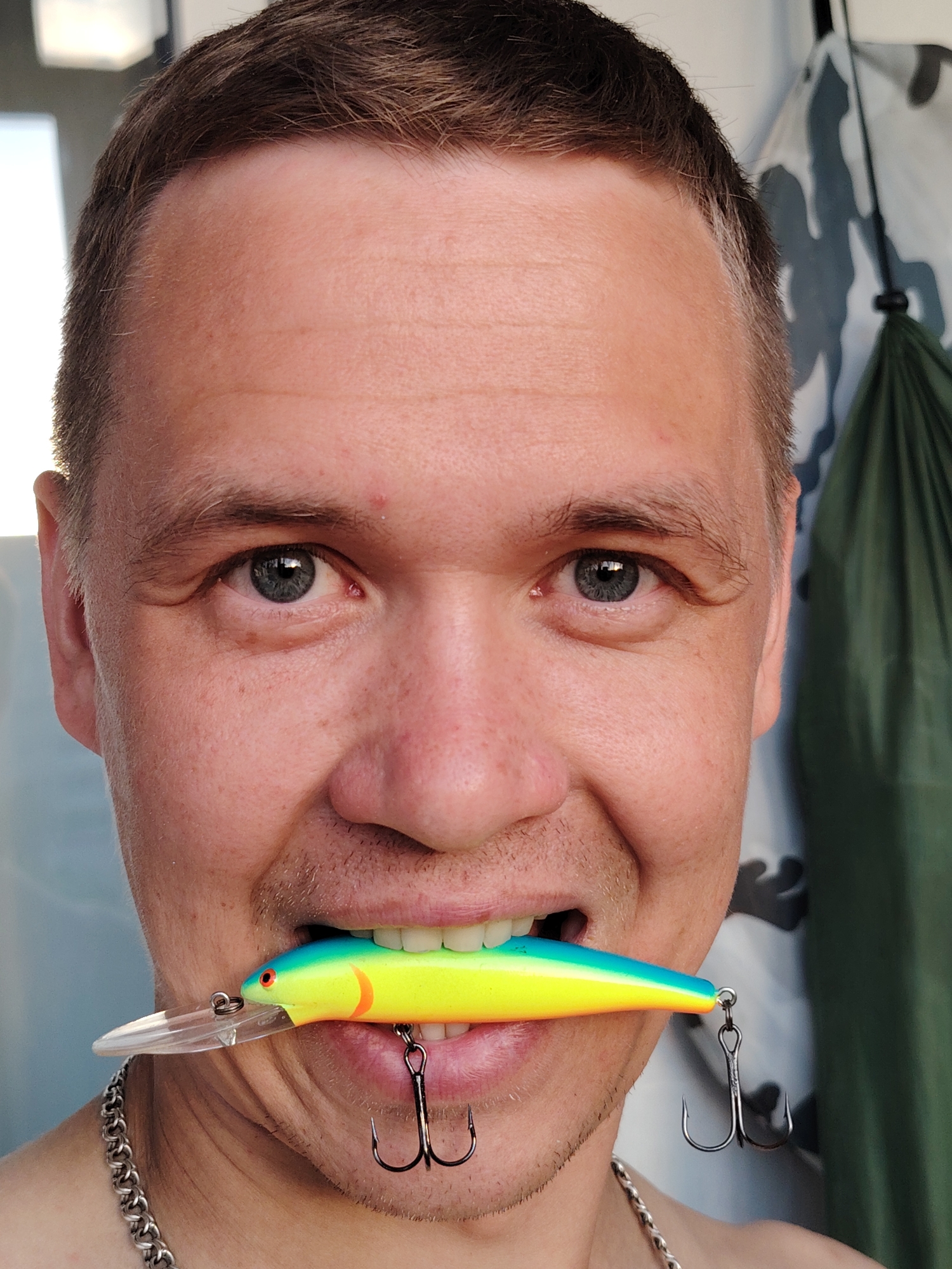 A close-up shot depicts a man holding a colorful fishing lure in his mouth. The lure is blue, yellow, and orange, with visible treble hooks. The man has short brown hair, light eyes, and fair skin. He is wearing a silver chain necklace. The background includes a light-colored surface and a dark green cloth with a camouflage pattern. There is no specific information in the image to indicate it was taken in Chaikovsky, Russia.