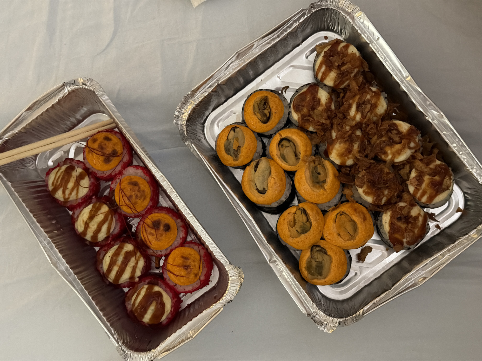 The image shows two foil containers of sushi rolls. The container on the left contains rolls with red rice and toppings in shades of orange and white with brown drizzling. A pair of chopsticks rests on top of the rolls in this container. The container on the right contains rolls with black seaweed and fillings in shades of orange and white with brown topping. The background is a wrinkled light gray surface. The scene is in Rossiyskiy, Russia.