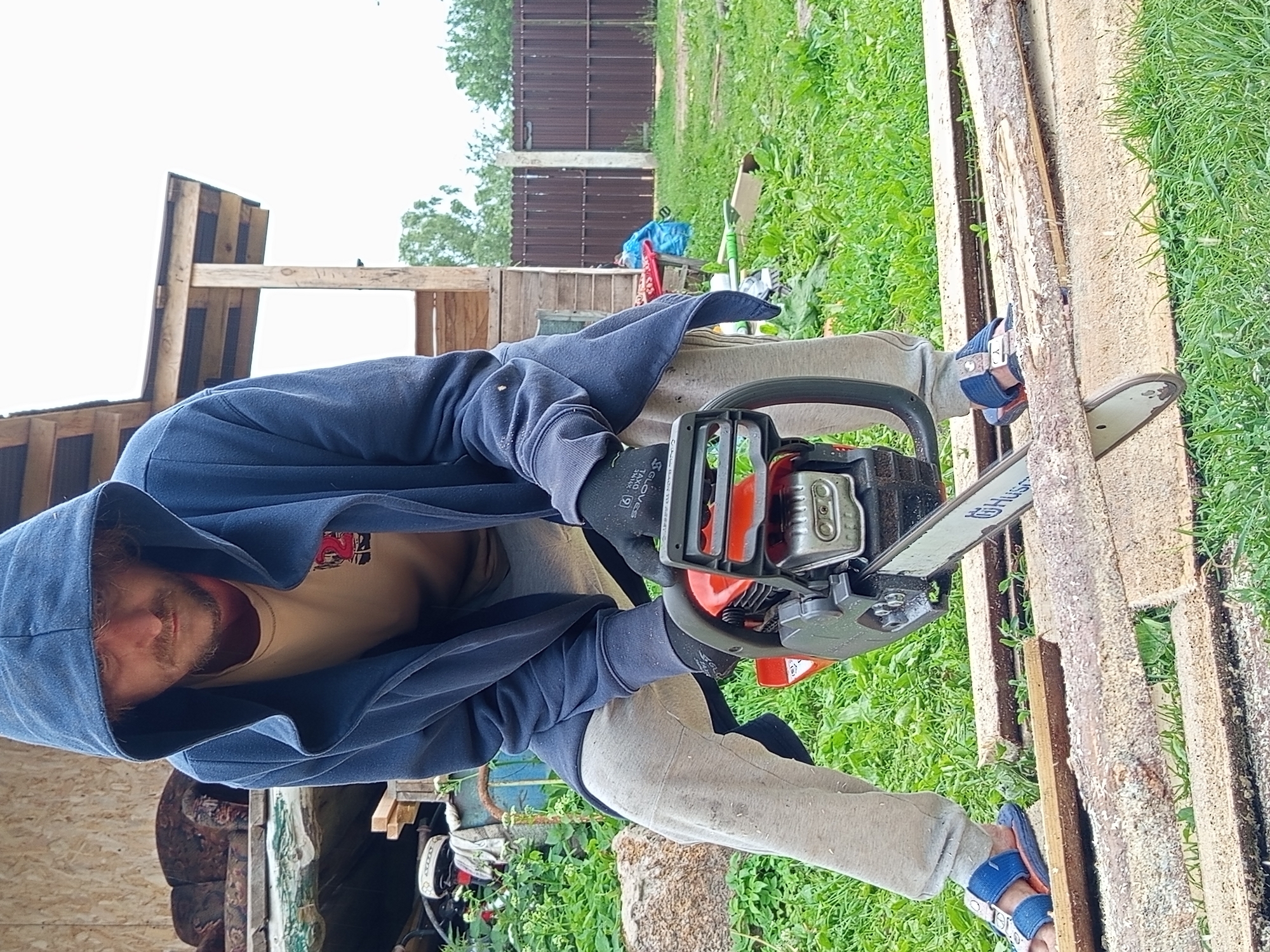 A man wearing a blue hoodie, grey sweatpants, and blue sandals is operating a chainsaw to cut wooden planks outdoors. He is wearing black gloves. The scene appears to be in a rural or suburban setting, possibly a backyard, with a wooden structure in the background and green grass covering the ground. There are several planks of wood on the ground, some of which have been partially cut. The man is positioned with his body angled towards the planks, holding the chainsaw with both hands and guiding it along a piece of wood. The overall impression is of a person engaged in a woodworking or construction task. This image was captured in Ostrovtsy, Russia.
