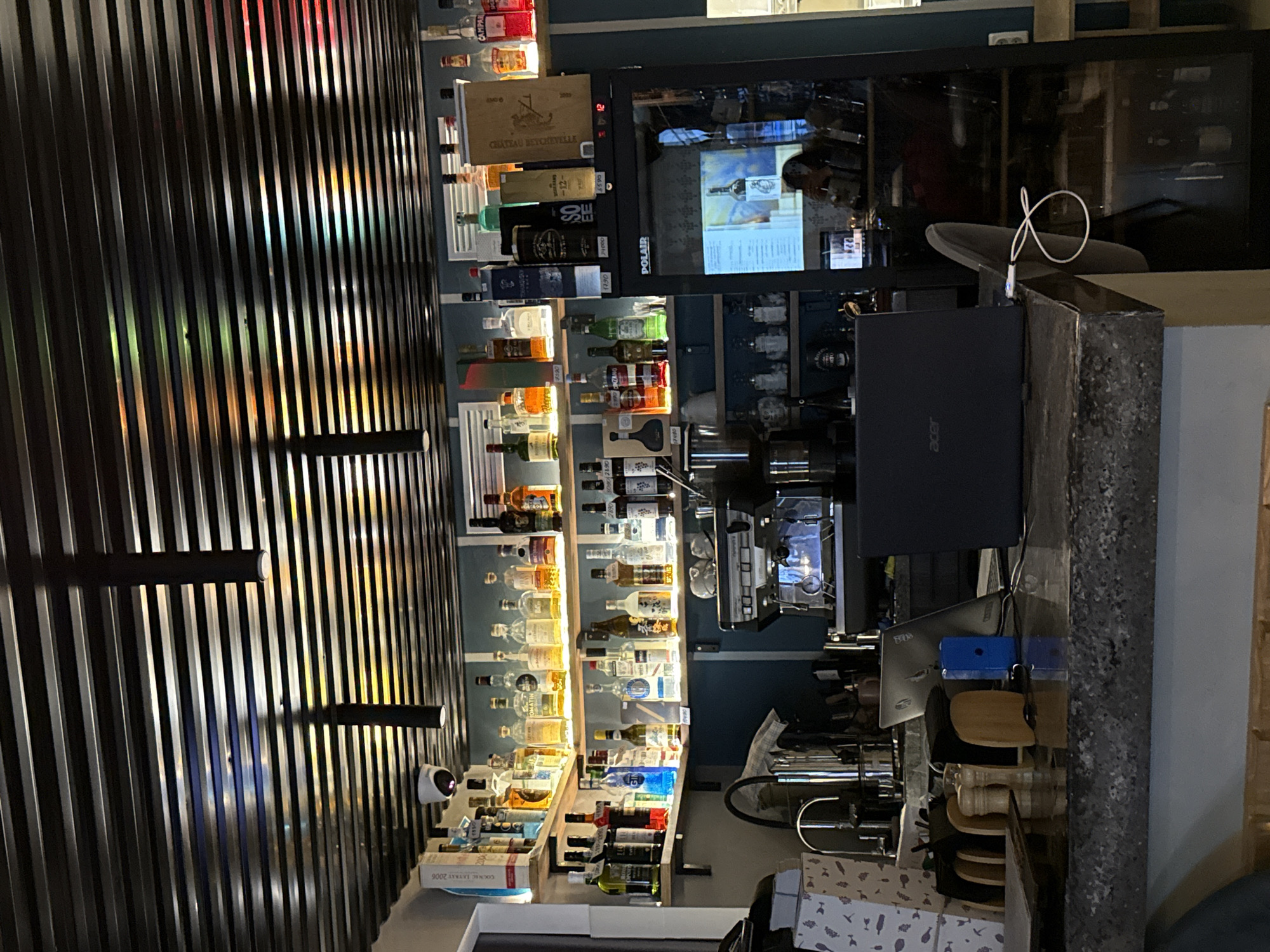 The image depicts the interior of a bar, likely in Izhevsk, Russia, with a focus on the shelves stocked with various bottles of alcohol. The ceiling features a modern, slatted design with hanging cylindrical lights and a security camera. The shelves are well-lit, illuminating rows of liquor bottles, some with labels visible like "Chateau Marceville" and "SO.EE." Below the shelves, a polished granite countertop runs across the foreground, adorned with a coffee machine, a laptop, various condiments, and a small blue object. A chair with a coiled cable is visible to the right, partially reflected in a glass-fronted cabinet displaying more bottles. The lighting is dim, creating a mood typical of a bar environment. There are no visible people actively engaged in service or consumption, suggesting the image was taken during off-hours or from a discreet vantage point.