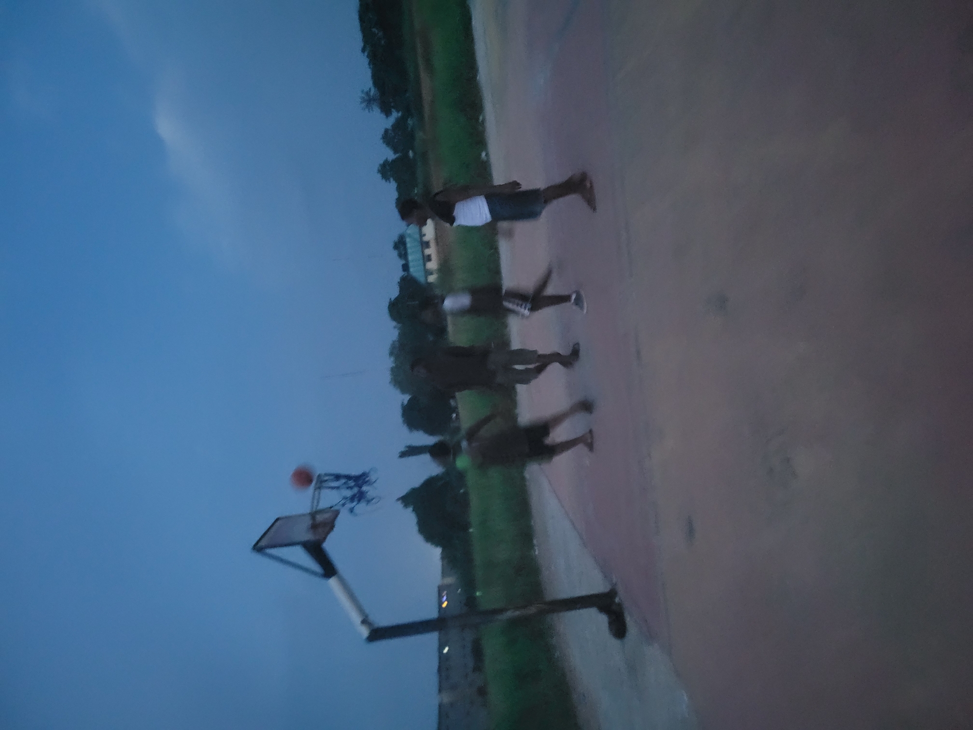 The image, taken in Egbagu, Nigeria, depicts a group of young people playing basketball on an outdoor court during dusk. The sky is a deep blue, indicating the transition from daylight to night. A basketball hoop with a net is visible, and a basketball is in the air, suggesting a game in progress or a shot being taken.
Three individuals are the main subjects. One person in the foreground appears to be in motion, possibly having just shot the ball or preparing to move. Another person is standing a bit further back, looking towards the hoop. A third person is partially visible behind them. The people are dressed casually, wearing athletic attire suitable for playing basketball.
The setting is an outdoor basketball court with a reddish-brown surface. To the left, a strip of green grass and some trees can be seen, with a building in the distance. The overall atmosphere is one of recreational activity and youthful energy. The dim lighting suggests the time is around evening, and the weather seems clear. There is no visible text in the image.