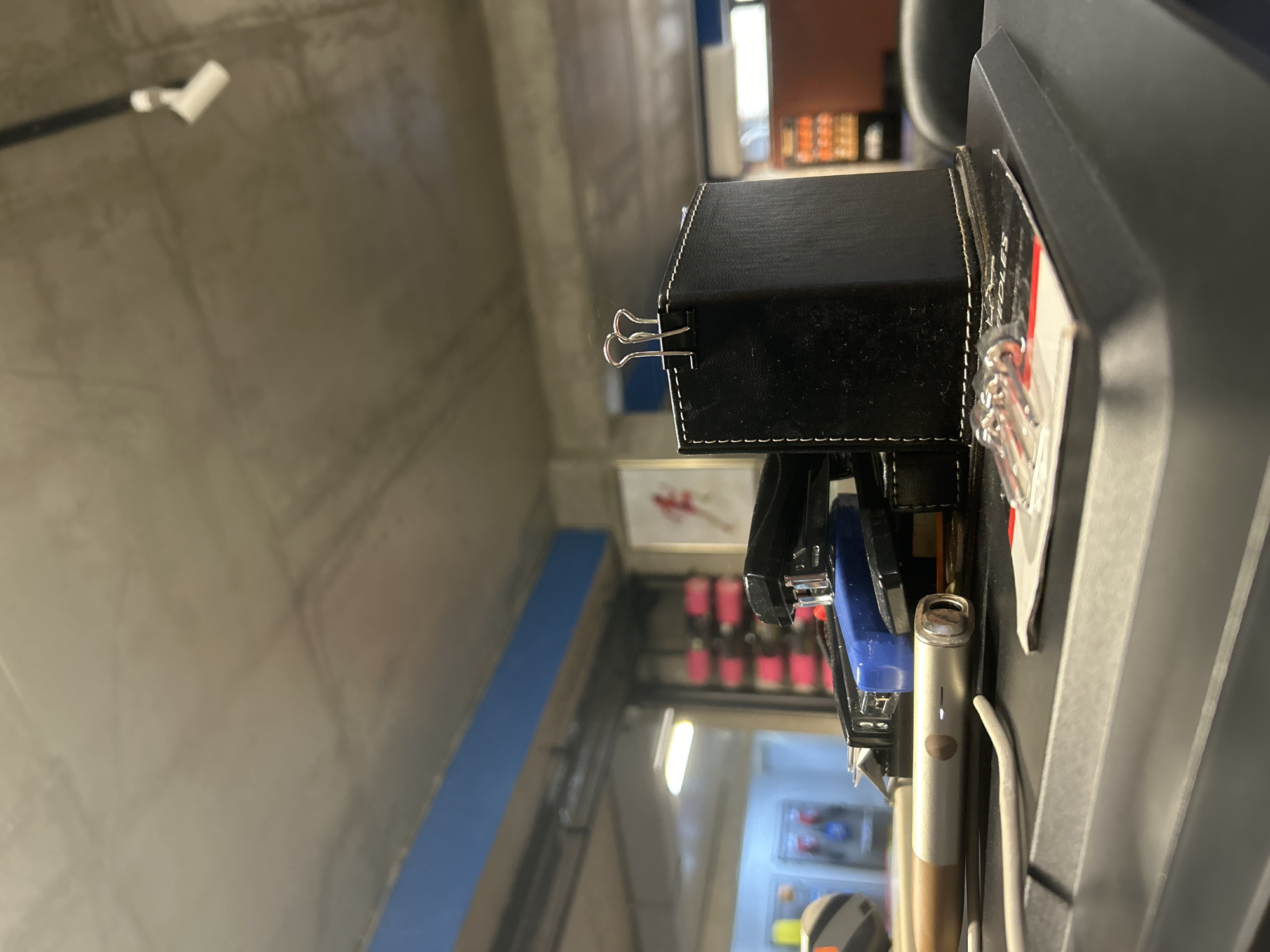 A close-up, low-angle shot captures a cluttered desk surface in an office environment, likely in Tbilisi, Georgia, given the context provided. The focus is on office supplies, including a black stapler stacked on top of a blue stapler, a small black leather pen holder adorned with white stitching and two silver binder clips, and a silver electronic device resembling a heating stick with a charging cable coiled nearby. To the right, a small packet labeled with red and white text, showing small metal needles, is visible. In the background, a concrete ceiling with exposed ductwork and a spotlight is apparent, along with blurry elements of office shelving and artwork on a distant wall. The overall impression is one of a functional, but untidy, workspace.