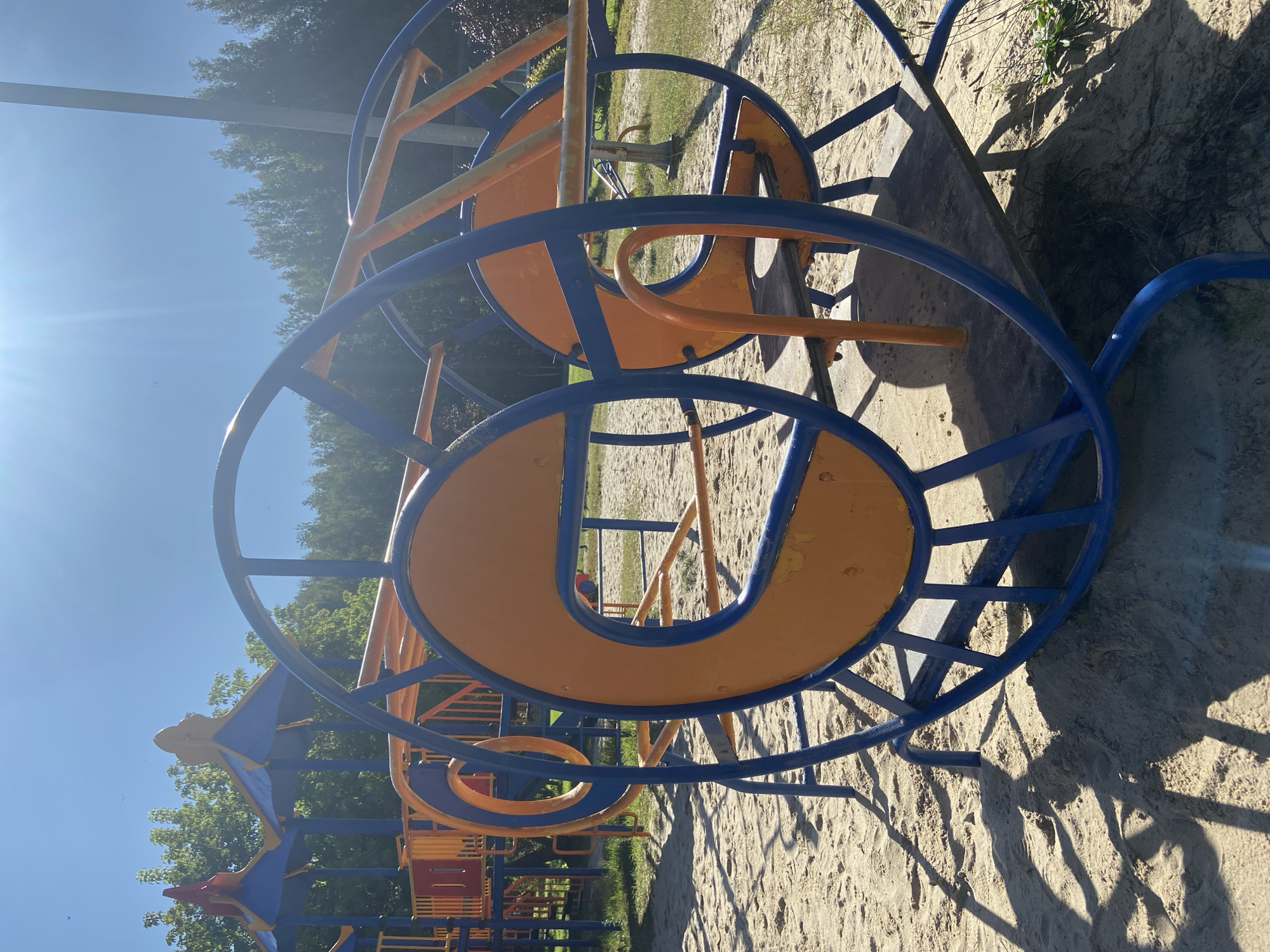 A blue and orange merry-go-round is in the foreground of a sandy playground. The merry-go-round is circular with a wooden seat in the middle. The merry-go-round is surrounded by sand and grass. In the background, there are trees and a blue sky with the sun shining. The playground equipment is colorful, with blue, orange, and yellow colors used throughout. The merry-go-round appears to be a piece of playground equipment designed for children to play on. The overall scene is of an outdoor playground on a sunny day. The location is specified as the city of Chulis, Russia.