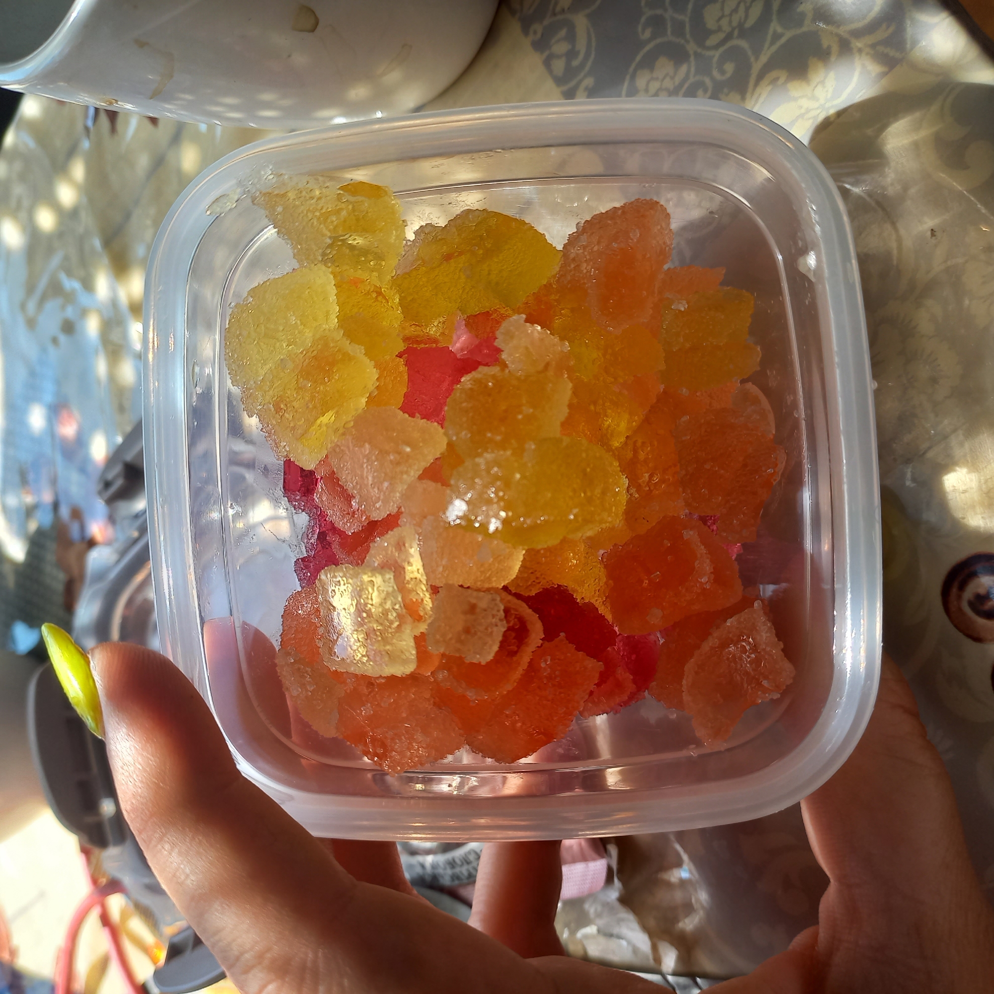 A person's thumb with a bright green nail is visible in the foreground, holding a clear plastic container filled with colorful, sugar-coated candies. The candies are in shades of orange, red, and yellow, and appear to be gummy or jelly-like. The container is partially filled, and the candies are piled high. The background is blurred, but suggests an outdoor setting with dappled sunlight. The image is taken from a close-up perspective, focusing on the candies and the hand holding the container. The specific location within Klyntsi, Ukraine, cannot be determined from the visual information provided.