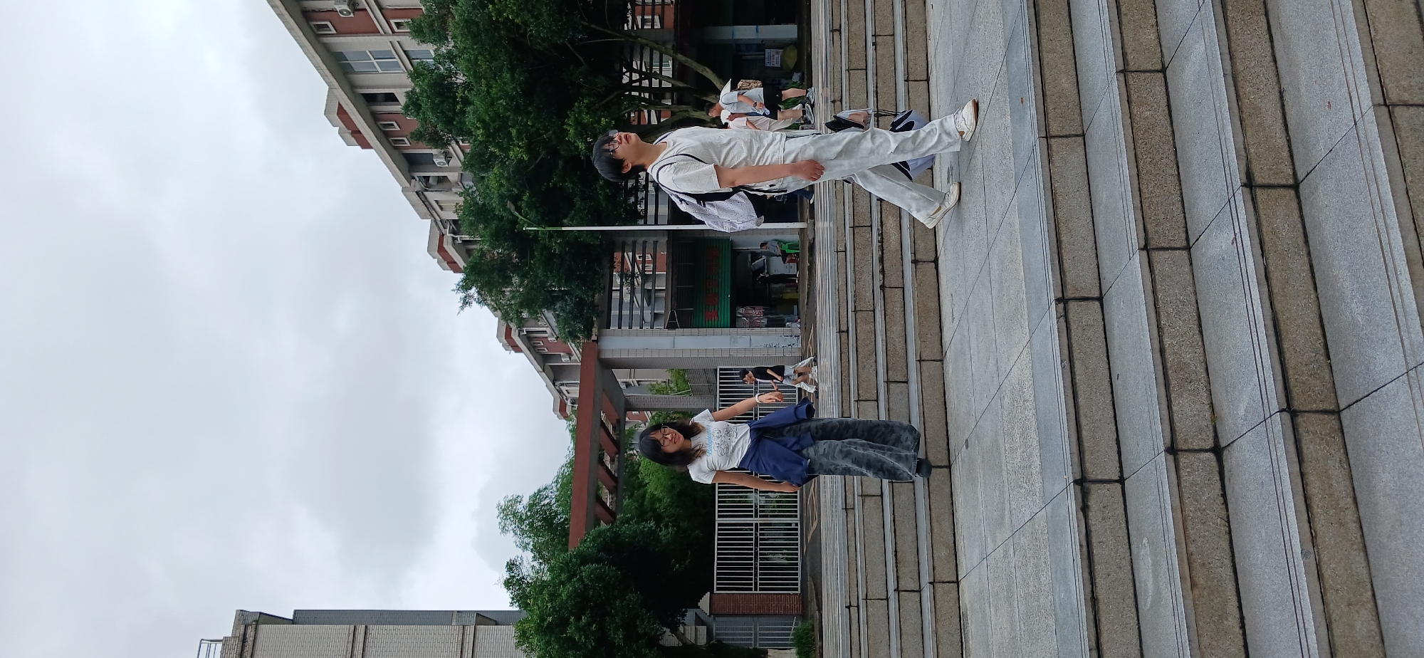 In Chongqing High-tech Zone, China, a wide staircase with stone treads and risers leads up to a building complex. The sky is overcast, with scattered clouds. On the stairs, a woman with dark hair, wearing glasses, a white t-shirt with a graphic print, a dark blue jacket tied around her waist, and camouflage-patterned wide-leg pants, walks down the stairs. To her right, a person with short dark hair, also wearing glasses, is walking up the stairs. This individual is wearing a white t-shirt and light-colored pants, and has a black and white backpack slung over their left shoulder. In the background, trees with dense green foliage are visible, interspersed with multi-story buildings. A metal fence borders the area to the left, and to the right, more buildings and some shops with signs are present. The overall scene depicts a casual outdoor setting on a cloudy day.