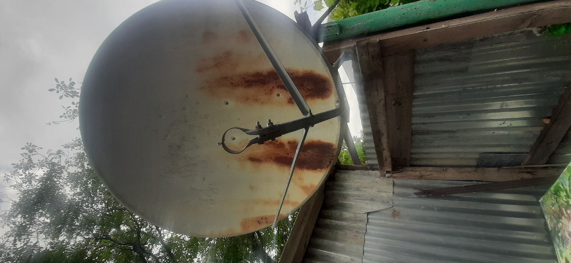 This image shows a satellite dish mounted on a rustic wooden structure. The satellite dish is large and off-white, with significant signs of rust and wear, indicating it has been exposed to the elements for some time. It is positioned at an upward angle, with the sky visible behind it. Surrounding the dish are the branches and leaves of trees, suggesting an outdoor setting.
The structure itself is composed of weathered wood beams and corrugated metal sheeting. Part of a green painted pipe is visible at the top, possibly part of a roof or support system. The overall impression of the scene is one of disrepair and age, with a functional but neglected satellite dish as the central object.
The weather appears to be overcast, with a muted sky that contributes to the somewhat somber mood of the image. The lighting is diffused, typical of a cloudy day. There are no people visible in the frame. The setting seems to be a rural or semi-rural area, possibly a backyard or an outbuilding, given the presence of trees and the utilitarian construction of the shelter. No text is visible in the image.