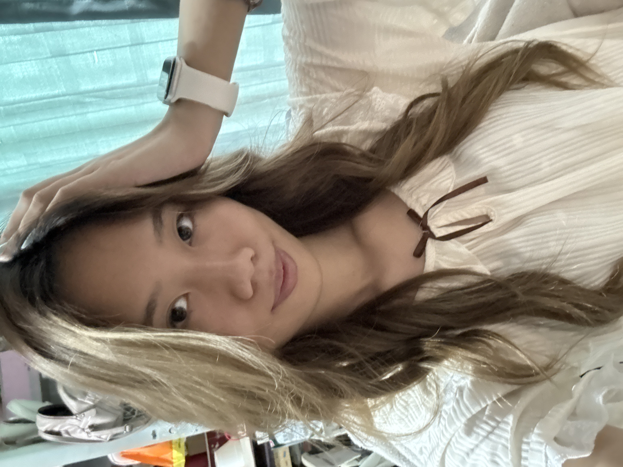 A woman is lying down, facing the camera. She is wearing a light-colored blouse with a ribbon tie at the neck and a white smartwatch. Her hair is light brown with blonde highlights. The background is blurred, showing a white textured surface and a glimpse of a teal-colored window or wall. The location is unknown and cannot be deduced from this image.