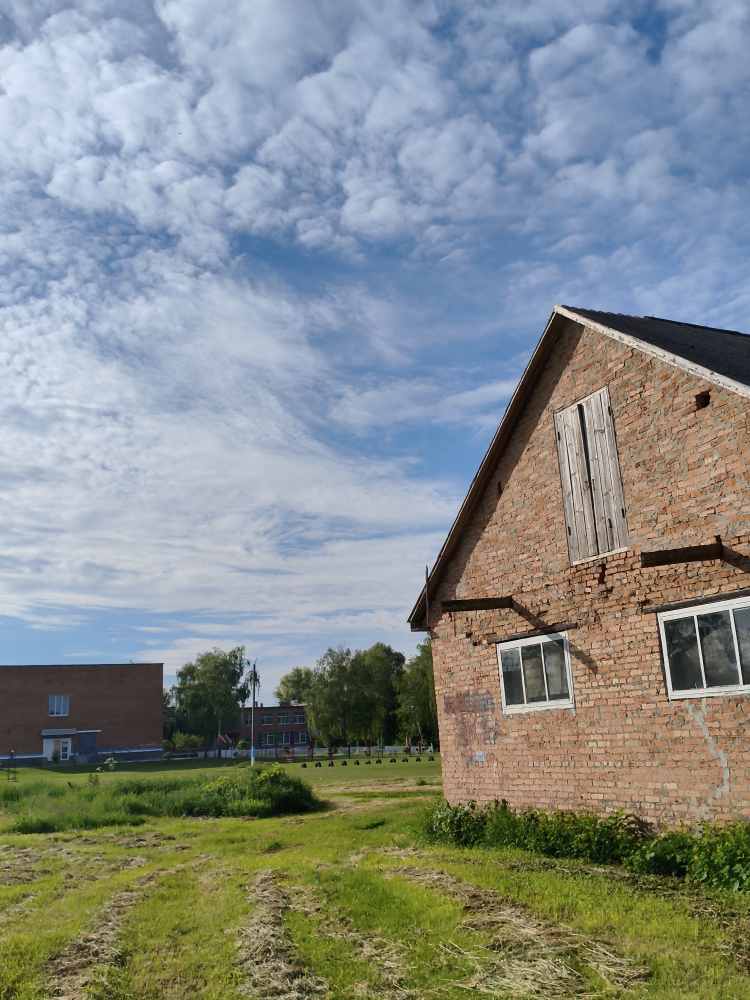 This media file presents a bright, serene landscape in Ustyvytsia, Ukraine. The central subject is a rustic, red brick building, possibly a barn or an older industrial structure, positioned prominently in the right foreground. It features a dark, sloped roof and an upper gable end with a pair of weathered wooden shutters. Several rectangular windows with white frames are visible along its side. The brickwork shows signs of age and repair, with some mortar lines and light-colored marks on the surface.
The immediate foreground and mid-ground are covered in vibrant green grass, interspersed with areas of lighter, possibly mown or dry, grass. A low line of bushes grows along the base of the brick building. Further back, a line of deciduous trees partially obscures other structures. To the left, a larger, more contemporary-looking brick building with distinct windows is visible, suggesting a school or public facility. Beyond the trees, glimpses of playground equipment can be discerned.
The setting is characterized by open space and a mix of natural elements and human-made structures. The sky is a clear blue, filled with a dynamic array of white clouds, ranging from fluffy cumulus to delicate cirrus formations, indicating a pleasant, sunny day, likely during late morning or afternoon. No specific activity or event is taking place, and no people are visible, contributing to a quiet and still atmosphere. There is no visible text in the image.