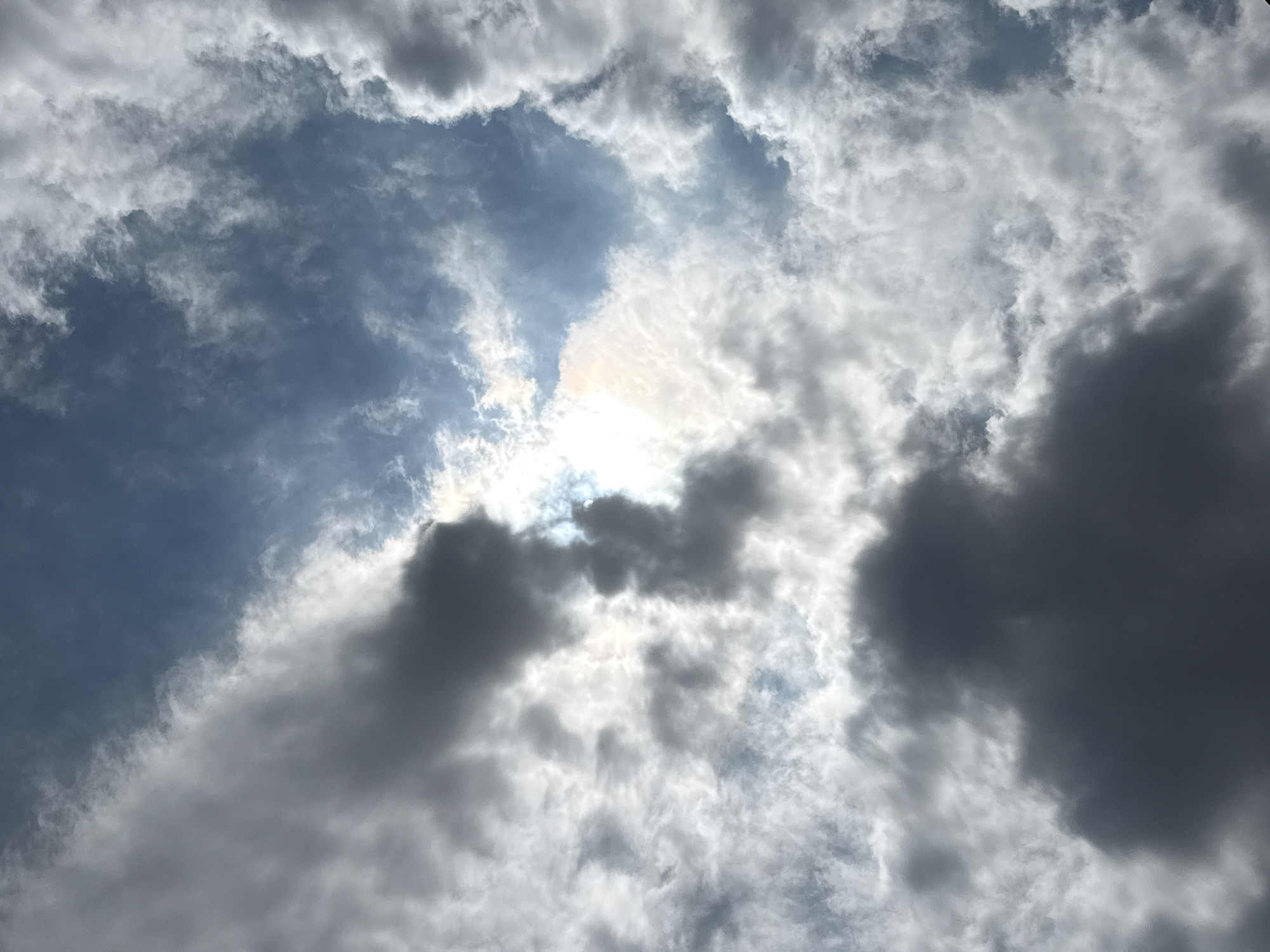 The image is a low-angle shot of a partly cloudy sky with the sun peeking through. The clouds are a mix of dark gray and bright white, creating a dramatic contrast against the blue sky. The sun is positioned in the upper center of the frame, partially obscured by a large white cloud, casting a bright glow. The scene appears to be an outdoor environment. There are no people, objects, or actions depicted in the image, and no identifiable landmarks or features that would indicate a specific location within Tbilisi, Georgia.