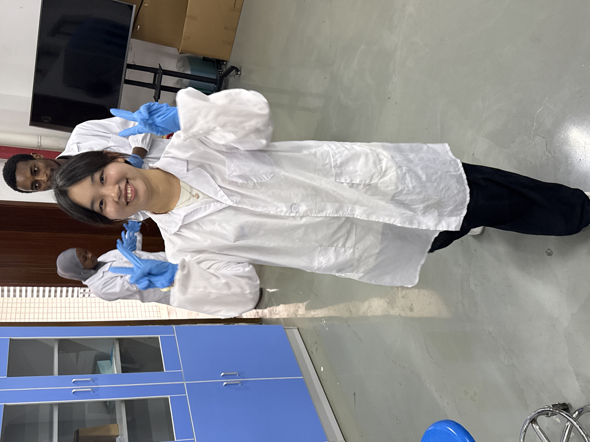 Scene: Interior, potentially a laboratory in Jiangjin, China.
People: A young woman is the primary subject, smiling and making a peace sign with both hands. She is wearing a white lab coat and blue gloves. Two other people are partially visible behind her, also wearing lab coats and gloves. One appears to be a man of African descent.
Subjects: The people mentioned above are the main subjects.
Objects: Blue laboratory cabinets are visible in the background. A television is mounted on the wall, and a cardboard box is stacked nearby. A blue stool and a metallic wheeled object are in the foreground. Lab coats and gloves.
Actions: The woman is posing for the camera. The individuals behind her are likely working or observing.
Interactions: The woman is the focus, potentially interacting with the photographer or others present but outside the frame.
Environment: The setting appears to be a laboratory or a room associated with scientific work, based on the presence of lab coats, gloves, and the cabinets. The floor is light-colored.