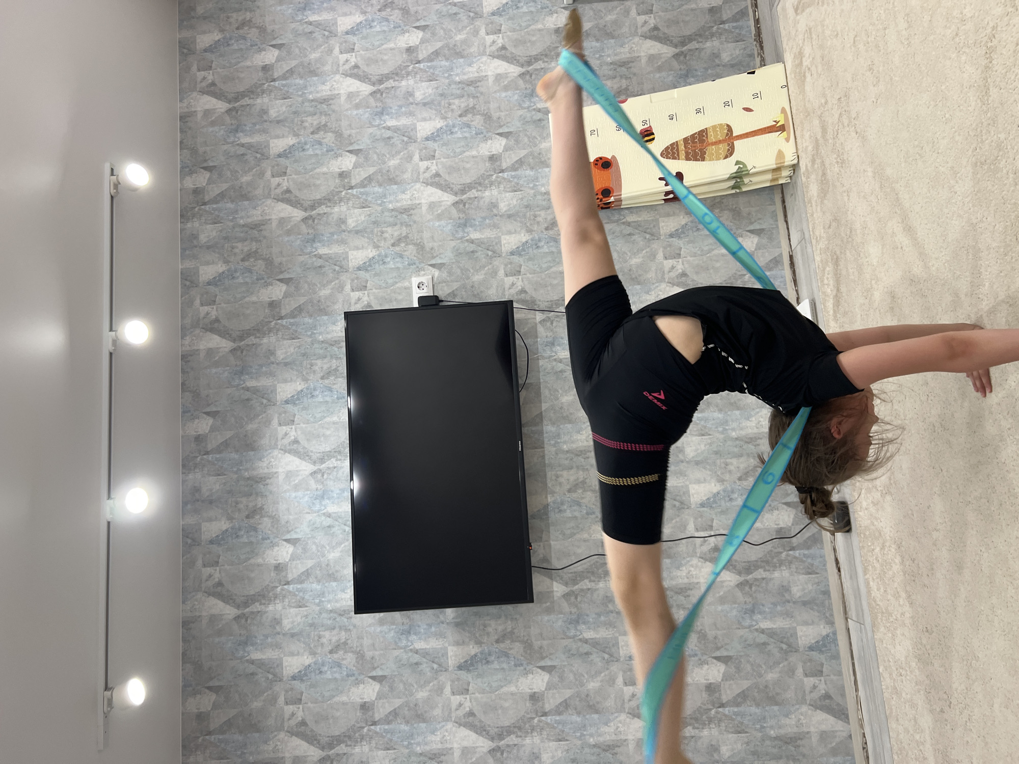 A girl in a black shirt and black shorts is performing a gymnastic split in an indoor environment in Chaikovsky, Russia. She uses a light blue resistance band. One leg is extended straight up, and the other is stretched forward. The girl is bending backwards so that her hands touch the floor, while keeping the legs straight in a split position. A television is mounted on a wall with patterned wallpaper. A standing lamp is visible on the left. A play mat is nearby.