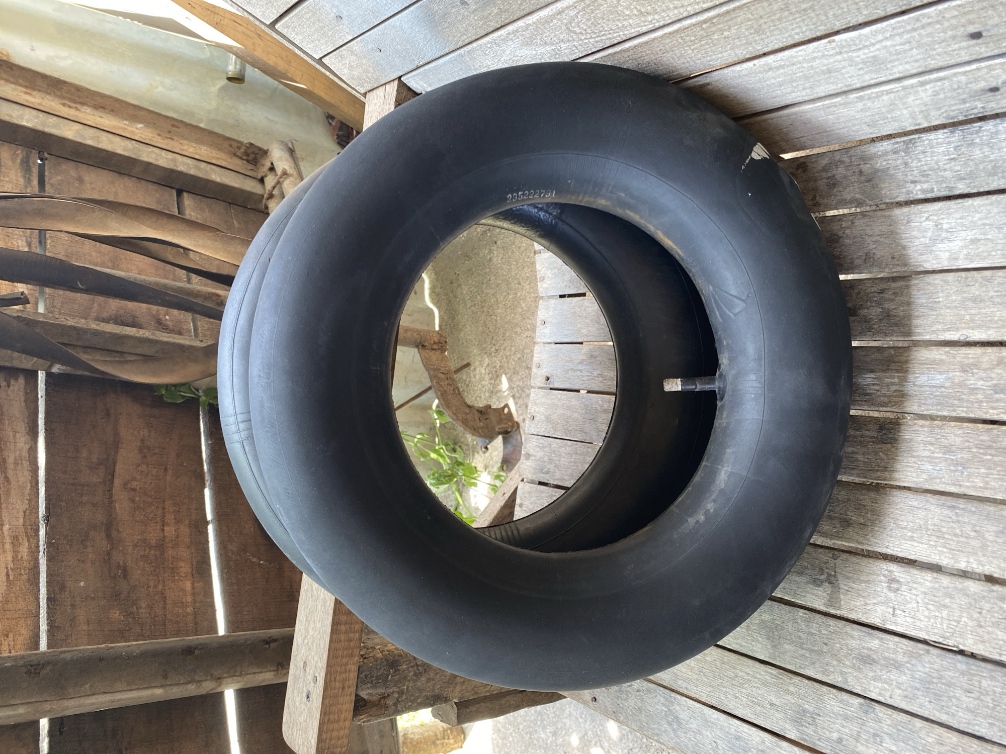 An inner tube lies on a rustic wooden surface, possibly a bench or table, in an outdoor setting. The background consists of weathered wooden planks and posts, suggesting an old structure or a rustic area. The inner tube is black and appears to be made of rubber, with a valve stem visible. The lighting suggests it is daytime, with shadows indicating sunlight. The specific location in Bayu, Indonesia, is not discernible from the visual information provided.