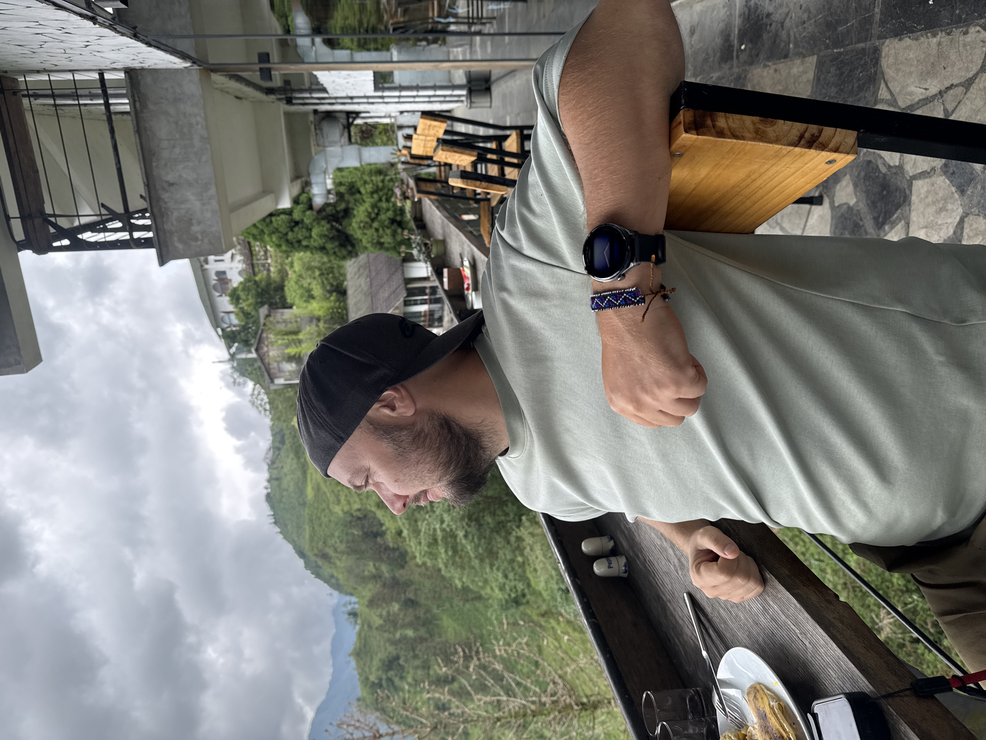 A man with a beard and a black baseball cap is seated at a table outdoors in Sa Pa Ward, Vietnam. He is wearing a light green t-shirt, a smartwatch, and a beaded bracelet on his left wrist. He is looking down towards his food. On the table, there is a plate with a meal, silverware, salt and pepper shakers, and some glasses. The background features a lush green mountainous landscape partially obscured by a cloudy sky. The architecture of the surrounding buildings suggests a mountainous tourist location. Several wooden chairs and tables can be seen in the background.