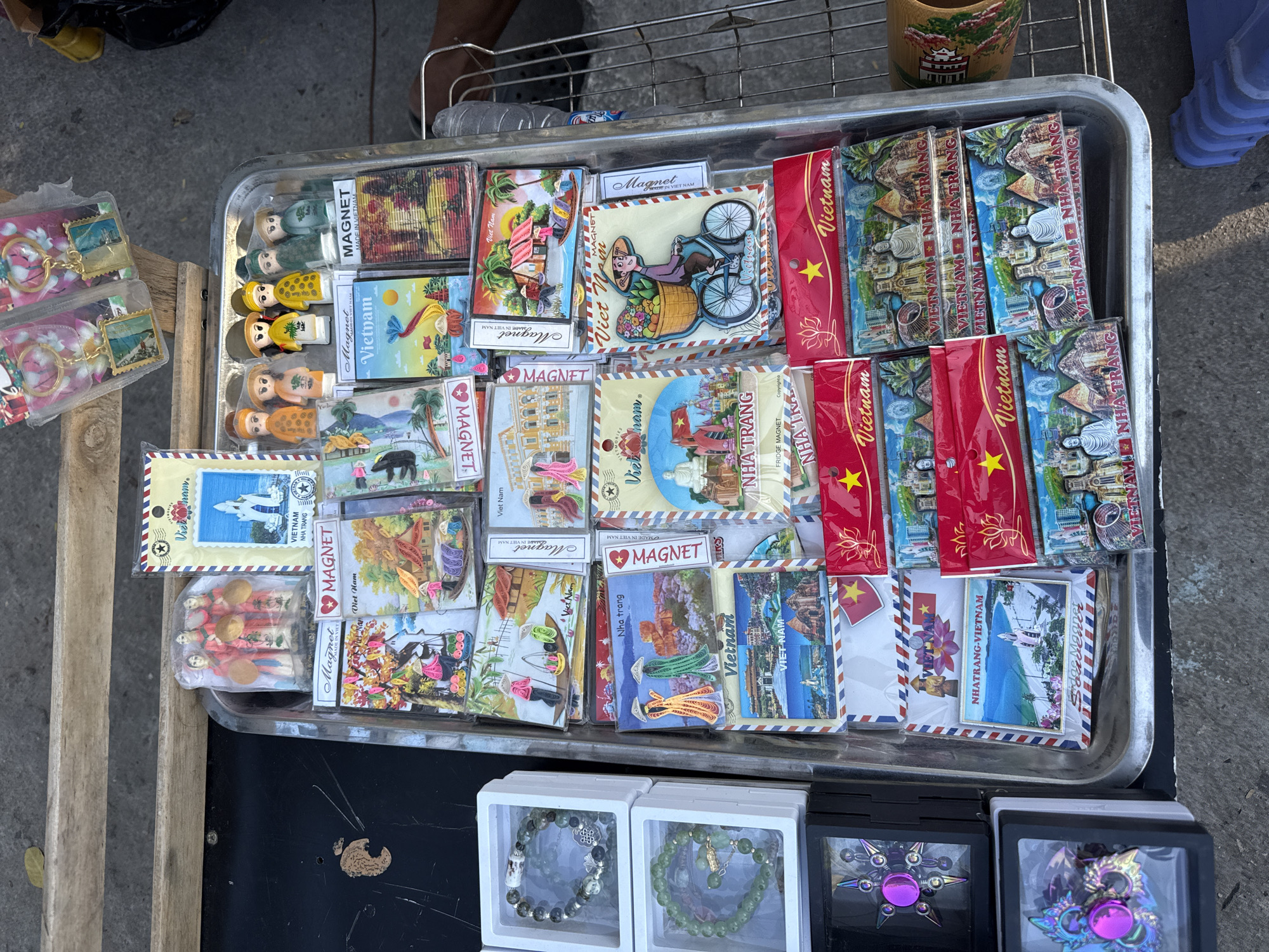 This image shows a street vendor's tray filled with souvenir magnets and other small items, likely in the city of Nha Trang, Vietnam. The magnets feature various scenes of Vietnam, including landscapes, landmarks, and cultural elements. Several red magnets with a gold star and the word "Vietnam" are also visible. Alongside the magnets, there are small figurines, keychains, and enclosed boxes containing beaded bracelets and spinners. The tray rests on a black surface near wooden planks and a section of a wire rack. The scene appears to be outdoors, with a portion of the ground and feet visible in the background.