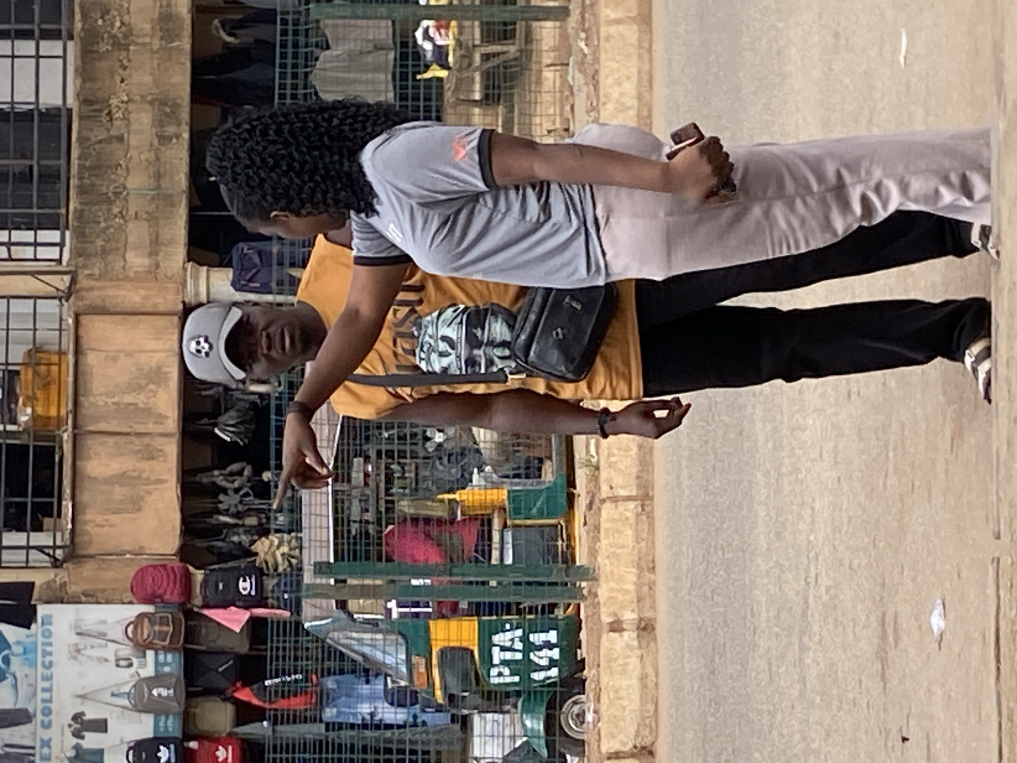 The image, rotated 90 degrees counter-clockwise, depicts an outdoor scene in what appears to be a bustling market or roadside area, consistent with the Bukuru, Nigeria location context.
Two individuals are the primary subjects. In the foreground, a man wears a yellow t-shirt and a grey baseball cap with a white emblem on the front. He has dark hair and a beard, and he is pointing downwards with his right hand, looking towards the left side of the frame. He is wearing a black shoulder bag. Behind him stands a woman with dark, curly braided hair, dressed in a grey short-sleeved t-shirt with an emblem on the sleeve, and light-colored trousers. She is holding a reddish-brown object, possibly a phone or wallet, in her right hand. Both individuals appear to be engaged in an interaction, possibly discussing items or directions.
The background features a market stall or shop. Immediately behind the man, another bag with a camouflage pattern is visible. The stall displays numerous bags of various sizes and colors, including red, black, brown, green, and blue, arranged on metal shelves or grates. To the left, part of a multi-story building with barred windows is visible. Further into the background, a small green and yellow vehicle, possibly an auto-rickshaw or Keke NAPEP, is partially visible.
Visible text includes "PTA 141" clearly printed on the side of the green and yellow vehicle. A sign on the far left, partially obscured, shows "EX COLLECTION". The lighting suggests it is daytime, with no strong shadows indicating a specific time of day. The scene conveys an everyday street or market environment.