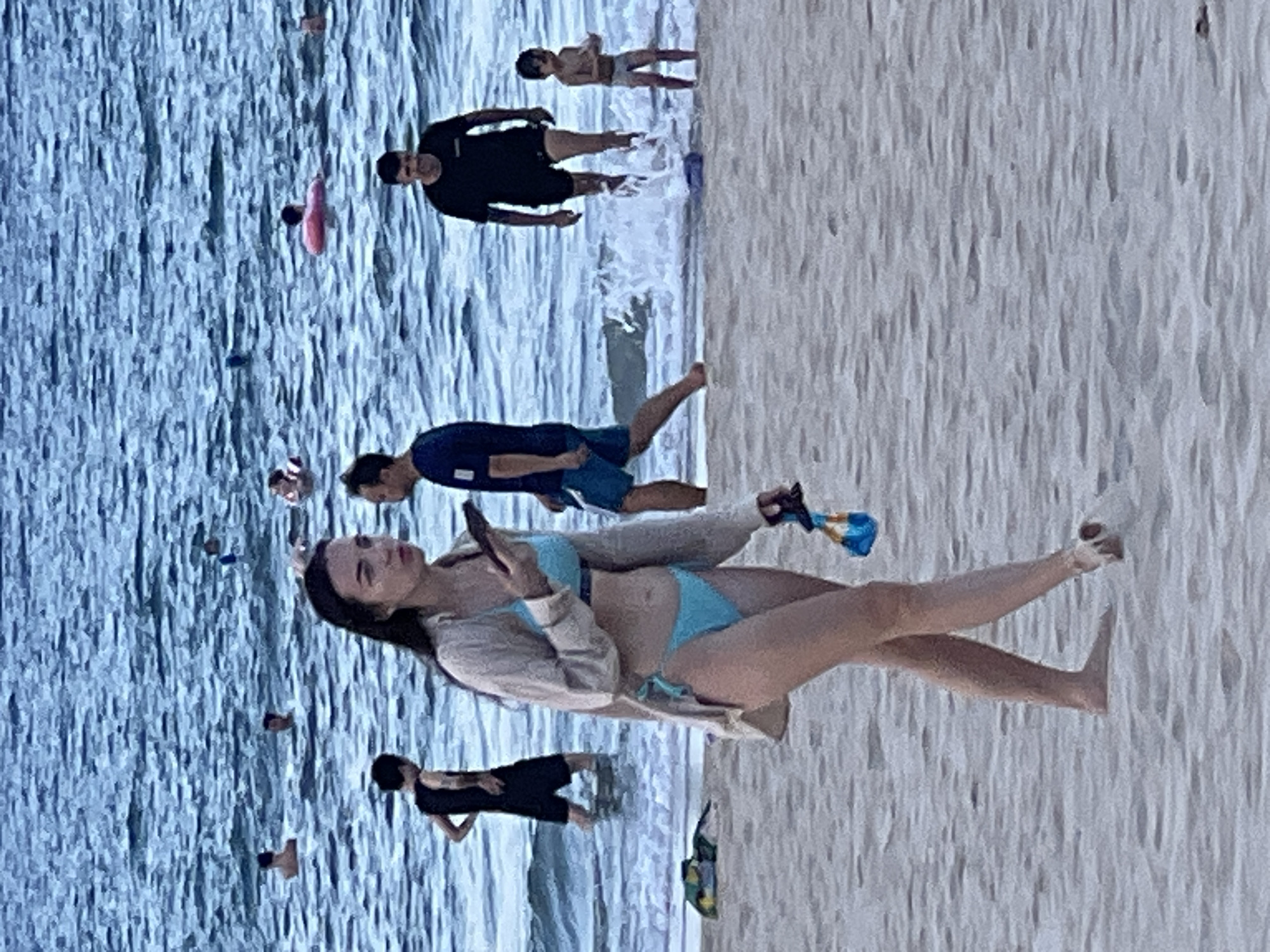 This image captures a scene on a sandy beach in Da Nang, Vietnam, with the ocean in the background. In the foreground, a woman wearing a light-colored overshirt and a blue bikini walks across the sand, holding a smartphone in her right hand and a small blue object with a black strap in her left. Several other individuals are visible in the water, some swimming and others standing. A young child stands at the edge of the water in the middle ground. The water is choppy, with small waves breaking near the shore. The overall lighting suggests it is daytime, possibly late afternoon or early evening.