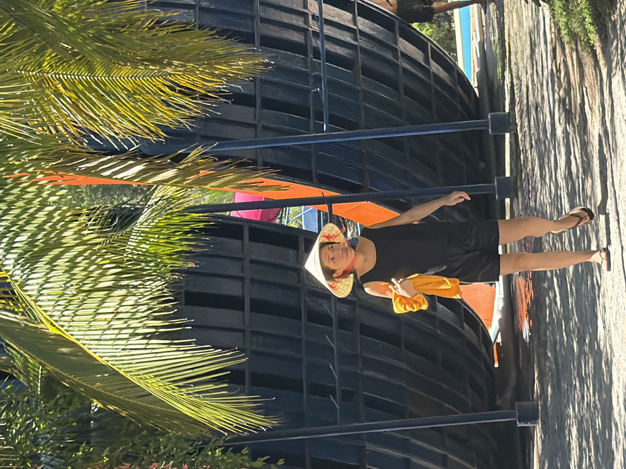 A woman wearing a conical hat and a black tank top walks on a paved surface in an outdoor area. She is holding a folded orange towel in her right hand. Behind her, a large, curved, dark structure with a grid pattern is visible, possibly part of an amusement park ride or attraction. Palm fronds hang from above, casting shadows on the ground. In the background, a blue swimming pool and another person are partially visible. The scene suggests a sunny day in a recreational setting, likely in Tay Nha Trang Ward, Vietnam, given the conical hat and tropical vegetation.