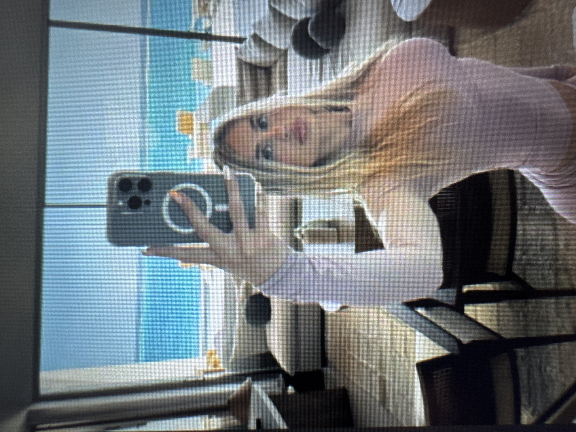 A mirror selfie captures a blonde woman holding a smartphone, likely an iPhone, in her right hand. The phone case features a visible MagSafe-like ring. The woman has long, straight blonde hair and is wearing a light pink, long-sleeved top or dress. Her nails are painted a light, possibly orange, color. She is looking directly at the phone's camera reflection with a neutral or slightly smiling expression.
The background, visible through what appears to be a large window or glass door, shows a clear blue body of water, strongly suggesting an ocean or a large pool, under a bright, possibly sunny sky. In front of the water, on an outdoor patio or balcony, there are several pieces of light-colored outdoor furniture, including chairs and sofas, and a railing. The interior space where the selfie is being taken also features some furniture, including dark-colored chairs. The scene suggests a daytime setting at a resort, hotel, or a high-end residence with an expansive water view. There is no discernible text visible in the image.