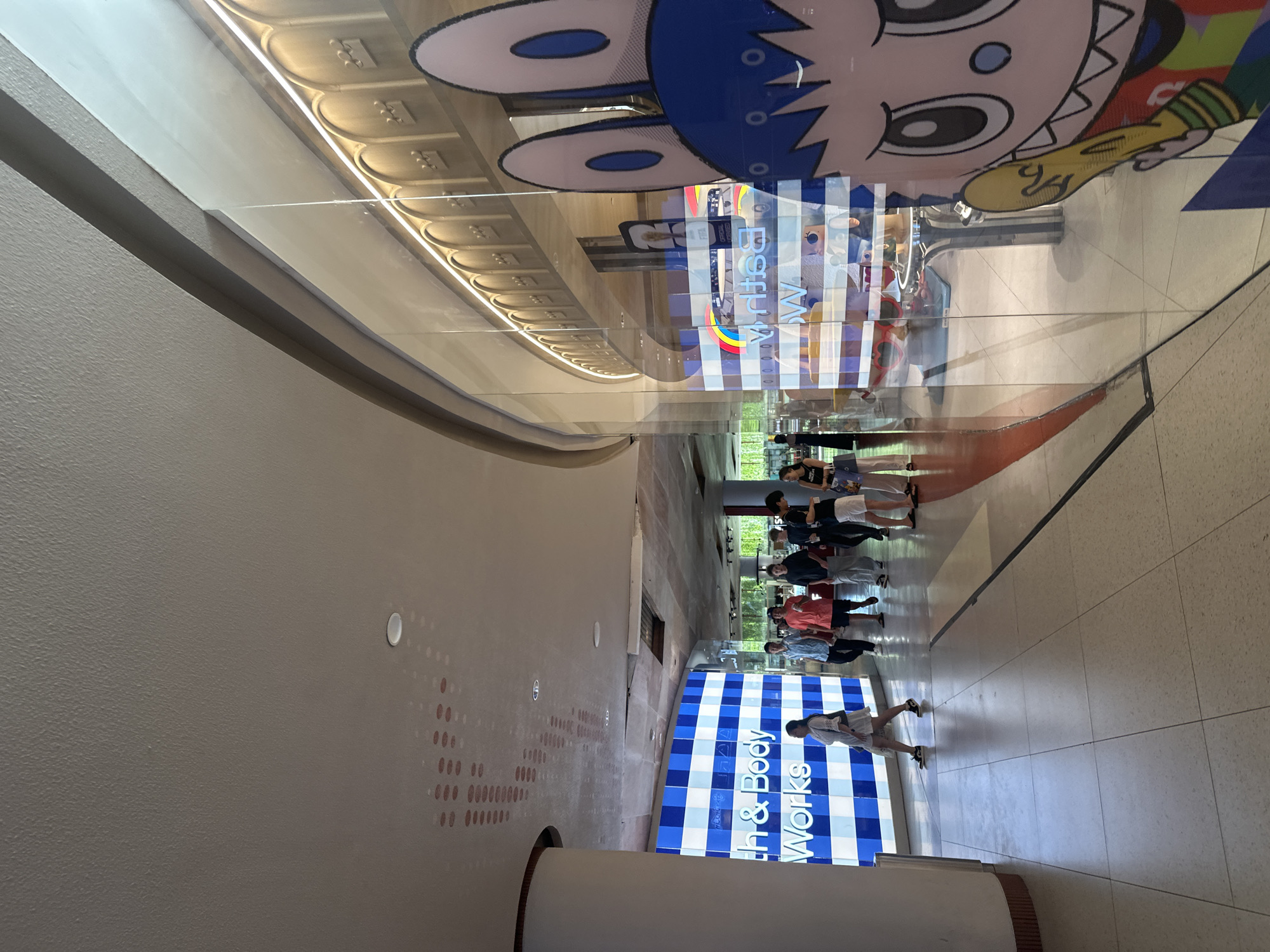 In Kuta, Indonesia, a wide shot captures a brightly lit indoor walkway, likely within a shopping mall. To the left, a large blue and white checkered display features the text "Bath & Body Works." Several individuals are walking in the corridor, some browsing and others in transit. The right side of the image is dominated by a glass storefront with a large, cartoonish character advertisement, featuring a character with blue hair and large ears, holding a yellow object. Reflected in the glass are more storefronts, a decorative arched wall with crown motifs, and an overhead light fixture. The floor is tiled with a light-colored, large square pattern. The ceiling is a light neutral color with some visible textures and light fixtures.