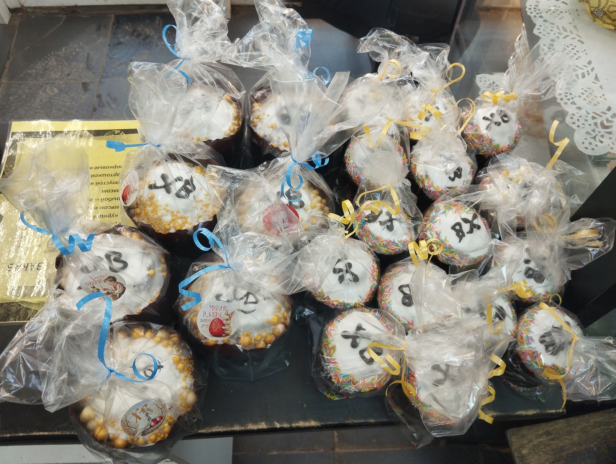 The image shows a display of individually packaged Easter cakes (kulich) on a dark surface, possibly a counter in a shop. The location is likely Votkinsk, Russia, based on the Cyrillic inscriptions on stickers attached to some of the cakes. The cakes are round with white icing on top, decorated with Cyrillic letters 'ХВ' (short for 'Христос Воскресе' - 'Christ is Risen') and colorful sprinkles or candied decorations around the edges. Each cake is wrapped in clear cellophane and tied with a ribbon, some with blue ribbons and others with yellow ribbons. Some of the cakes have small stickers with Easter-themed images or greetings in Russian. A yellow sign with Cyrillic text is partially visible on the left.