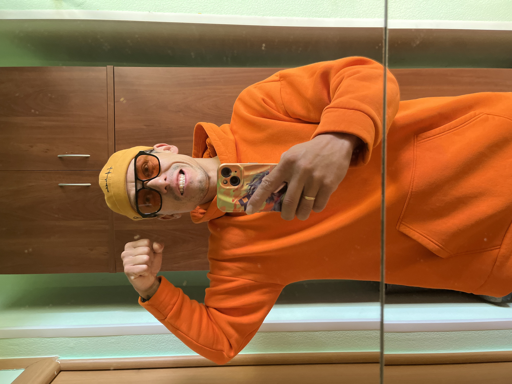 A man is captured in a mirror selfie, seen from the chest up, wearing a bright orange hooded sweatshirt, a yellow beanie, and orange-tinted sunglasses. The image is oriented 90 degrees clockwise. He smiles broadly with his teeth showing, looking directly at the camera.
In his right hand, he holds a smartphone with a distinctive, colorful case that features an illustration and multiple camera lenses. A gold-colored band ring is visible on his right ring finger. His left hand is clenched into a fist, positioned near his chest.
The background, reflected in the mirror, shows a wooden cabinet or wardrobe with a warm brown finish and silver horizontal handles. Above the cabinet, there is a light green, textured wall, separated by a white horizontal trim. A thin, vertical line, likely the edge of a mirror or frame, runs along the right side of the composition. The man's expression suggests happiness and confidence. The setting appears to be an indoor space with artificial lighting. Text on the yellow beanie is visible but illegible.