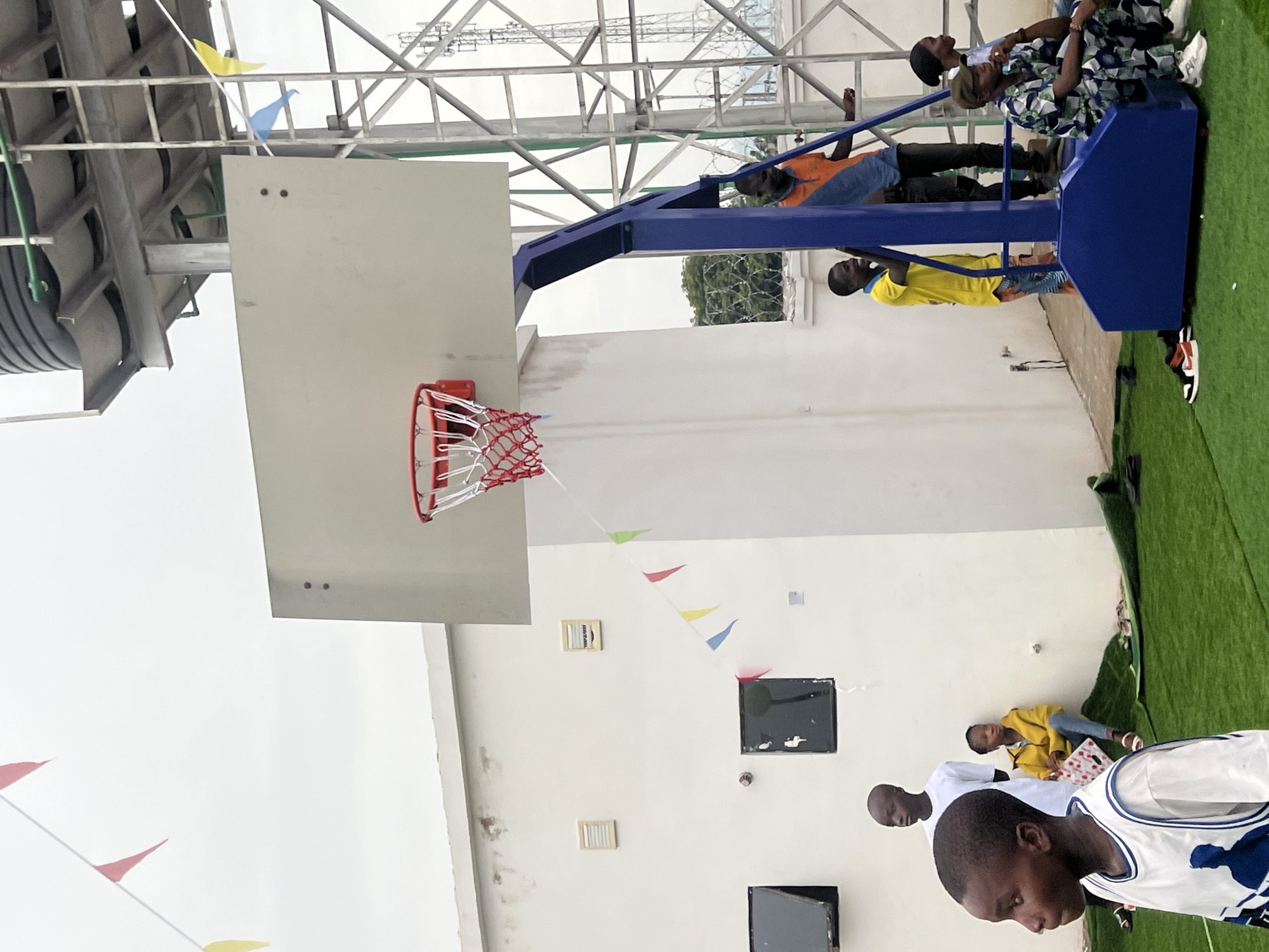 The image shows a partially constructed basketball court at an outdoor event in Howayi, Nigeria. A basketball hoop with a white backboard and a red and white net is visible. People are present; some are gathered near the base of the basketball stand, and others are on the stand itself. The environment includes a white wall with decorations, green artificial turf, and overhead metal structures. The weather appears overcast.