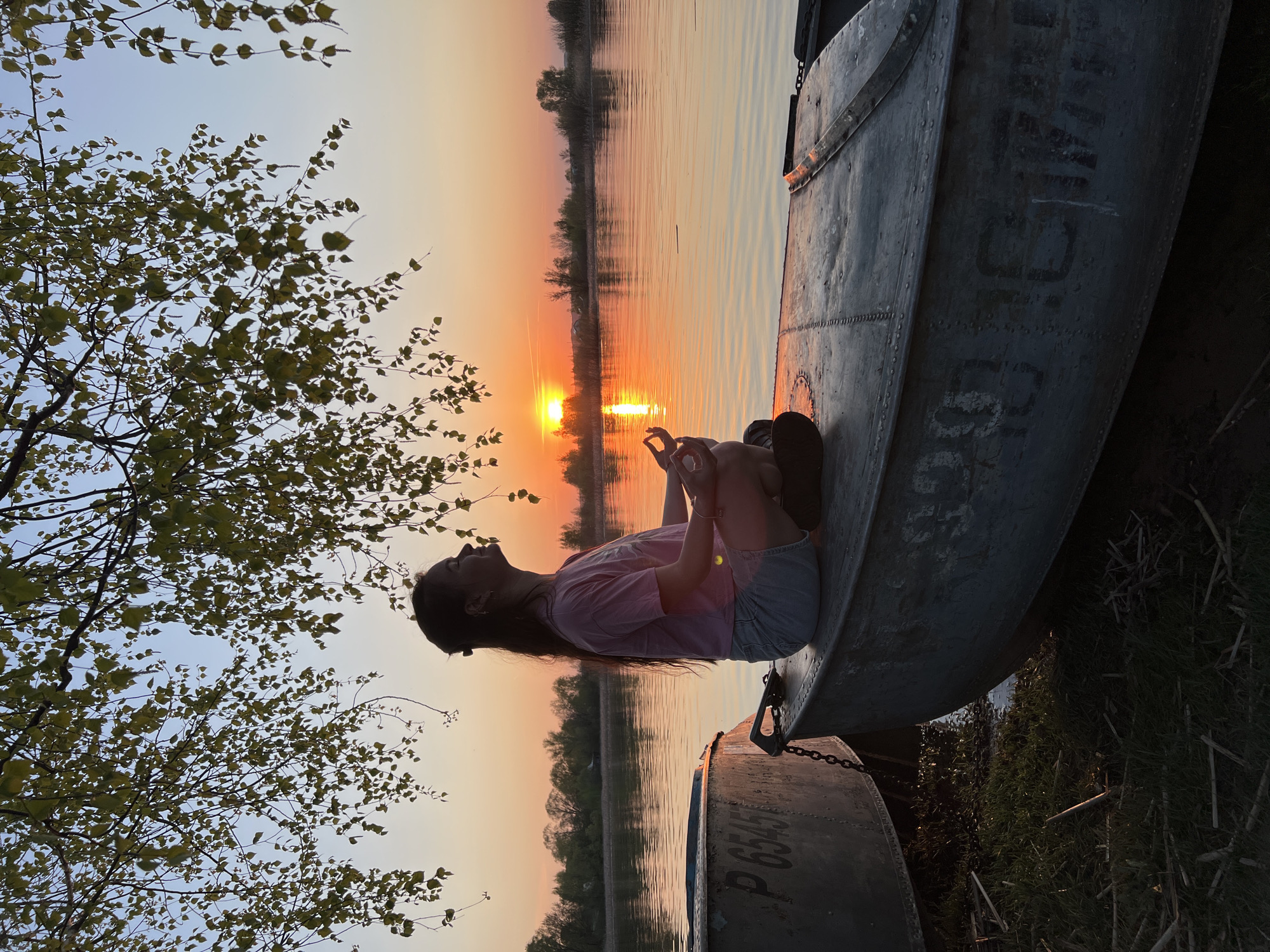 A long-haired woman is sitting in a metal boat at sunset. The location is most likely near Borovoye Matyushino, Russia. She's wearing a pink shirt and denim shorts. The sun is reflected on the water behind her. In the bottom right corner and bottom left corner is another boat. A tree is situated on the left-hand side.