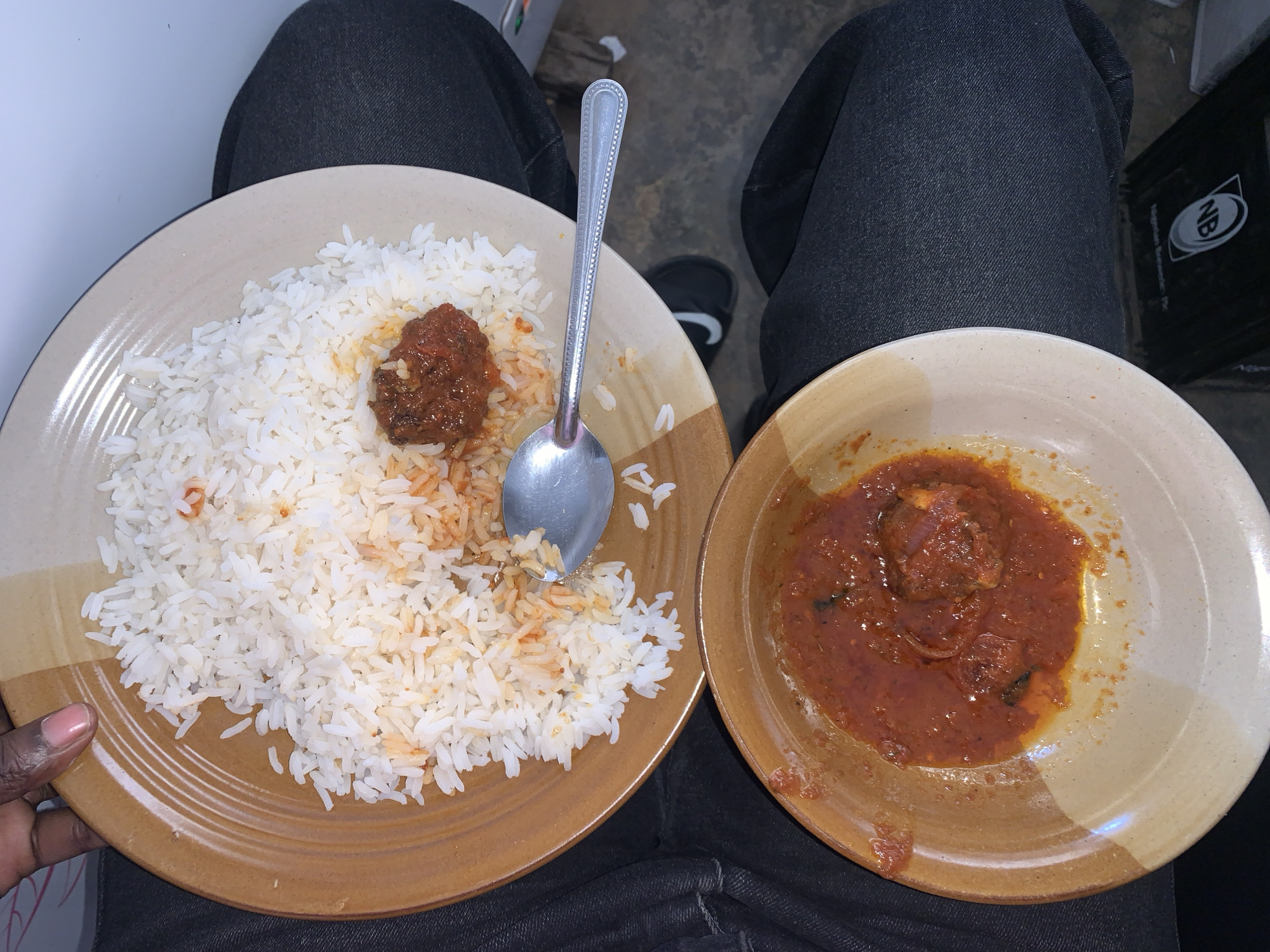 The image presents an overhead view of a casual meal set on a person's lap, likely indoors. The main subjects are two plates of food: a large plate of white rice and a smaller bowl of stew or sauce.
The larger plate, with light brown and off-white concentric bands, holds a substantial amount of white rice. Parts of the rice are mixed with a reddish-brown sauce, and a single, sizable piece of meat or similar ingredient rests prominently on top. A silver spoon with a decorative, ridged handle is placed on the rice. A hand with dark skin and a light pink fingernail is visible holding the edge of this plate on the left.
The smaller, deeper bowl to the right, matching the larger plate's design, contains a generous portion of thick, reddish-brown stew or sauce. Several pieces of meat, fish, or other chunky ingredients are submerged in the stew.
The person is wearing dark-wash denim jeans. On the right side, a black slide sandal with a white Nike swoosh logo is visible on the floor near the right leg. In the background, towards the upper right, a black bag or container is partially seen, featuring a white "NB" logo and the text "Nigerian Bottling Plc.", indicating the location is in Nigeria, consistent with the provided context of a "New Stadium" in Nigeria. The lighting suggests an indoor setting or a time of day with indirect light. The overall scene depicts a simple, hearty meal.