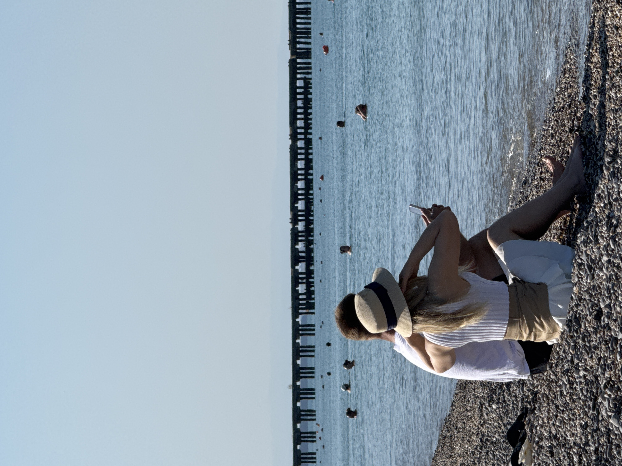 The image depicts a couple sitting on a pebble beach in Novofedorovka, Ukraine, facing the sea. The man is wearing a white t-shirt and dark shorts, and the woman is wearing a straw hat with a blue ribbon, a white ribbed tank top, and a light-colored skirt. The woman is holding a smartphone and appears to be taking a selfie. Several people are visible in the water, swimming and wading, with a long pier extending into the sea in the background. The sky is clear and pale blue, and the water is a calm blue. The shoreline is covered in smooth, gray pebbles.