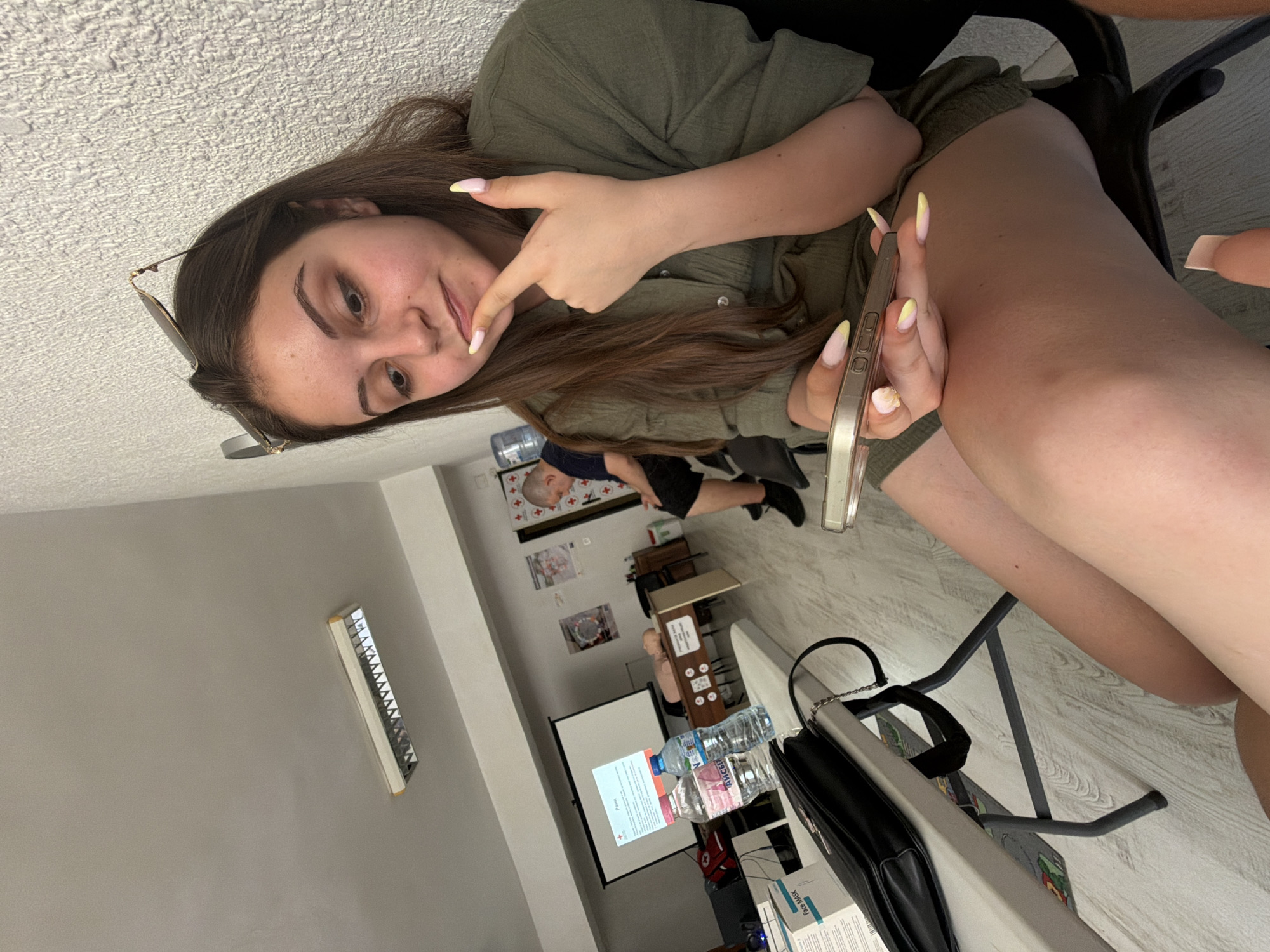 In Burgas, Bulgaria, a young woman with long brown hair, wearing a green dress and sunglasses perched on her head, sits at a table, holding a smartphone. Her nails are painted with a yellow and pink gradient. She is looking down at the phone, her finger touching her chin in a pensive gesture. To her left, on the table, are two water bottles and a black purse. In the background, a projector screen displays text. A mannequin used for medical training lies on a desk, and a man in a dark shirt and shorts is seated further in the background, facing away from the camera. The room appears to be a classroom or training facility, with fluorescent lighting on the ceiling and a textured wall.