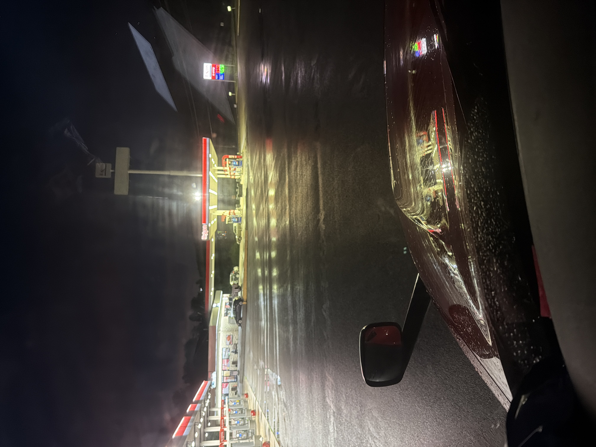 The image depicts a view from inside a vehicle at night, looking out towards a gas station. The ground is wet and reflects the lights of the gas station and nearby buildings. A red truck's hood is visible in the foreground, with raindrops on its surface. The gas station has red and white awnings covering the pumps, which are illuminated. A sign with "Exxon" is visible. There are several cars parked at the gas station, and a few indistinct figures are present. The sky is dark with some clouds. The scene appears to be a typical gas station illuminated at night, likely after or during rainfall, in the city of Wildwood, United States.