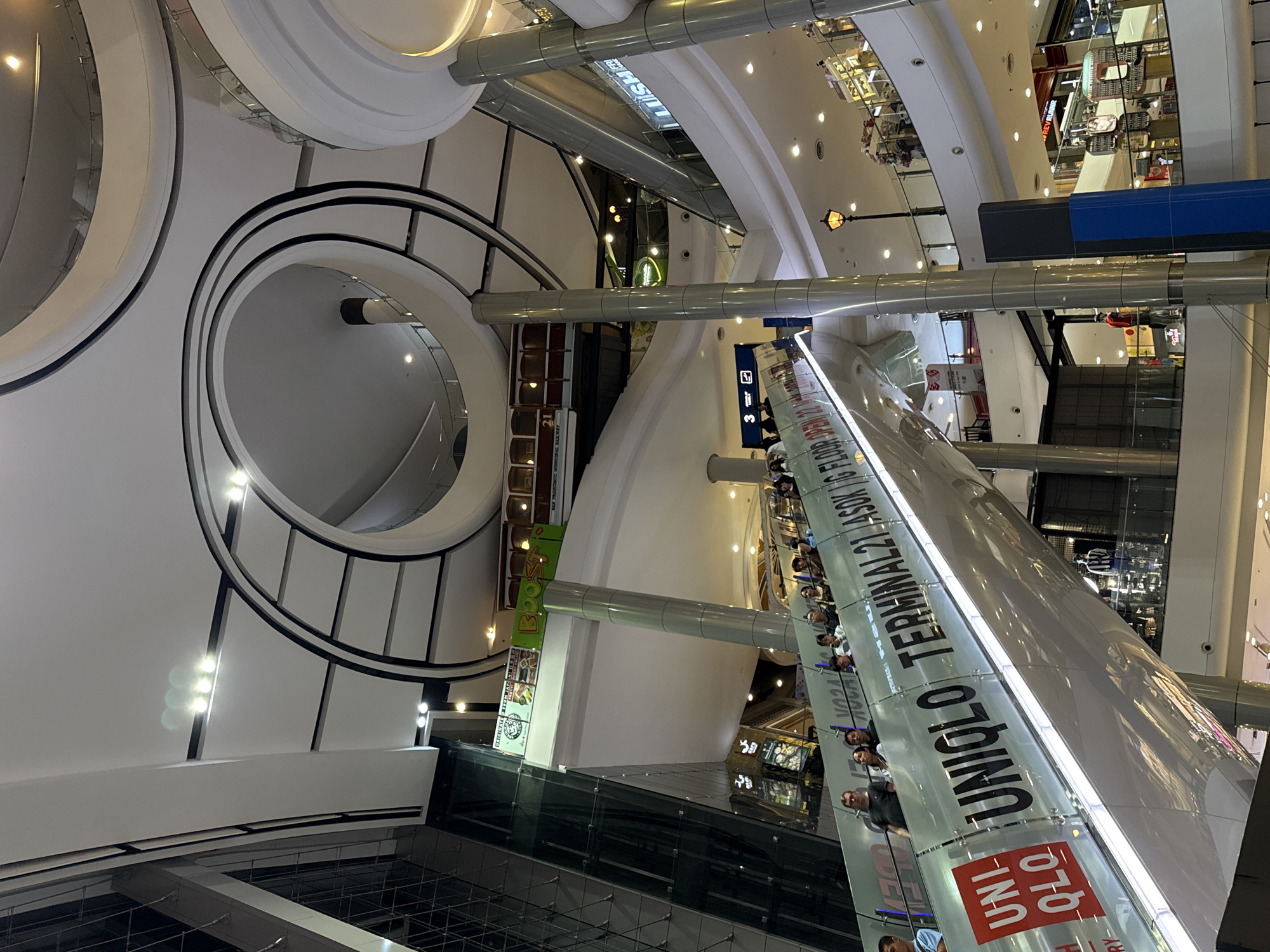 The image is a high-angle view inside a modern shopping mall in Bangkok, Thailand. A circular, multi-tiered architectural element dominates the left side of the frame. An escalator with advertising for Uniqlo and Terminal 21 Asok is prominently visible, and people are riding it. The mall has multiple levels, visible in the background, with a variety of stores and lighting fixtures. The environment is brightly lit and appears spacious.