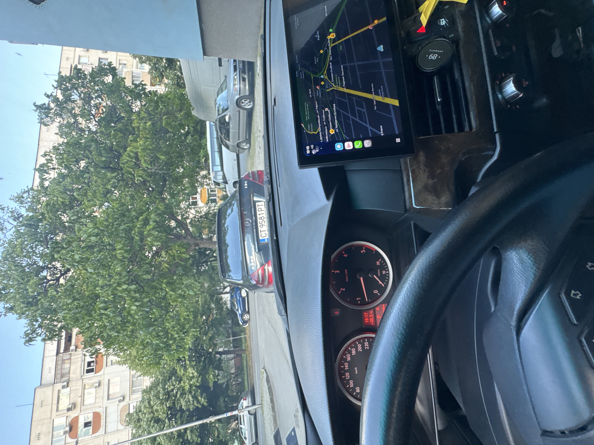 The image depicts the interior of a car, specifically the dashboard and steering wheel. A navigation system is visible, displaying a map of Stara Zagora, Bulgaria. Key locations shown on the screen include "бул. Цар Симеон Велики" and "ул. Сливница". The car's dashboard displays a speed of 0 km/h. Another car is stopped ahead, and its license plate reads "CT 9581 PT", indicating a Bulgarian license plate. The environment appears to be an urban street with trees and buildings in the background. The time on the instrument cluster is 19:17 and the temperature is +20.5 degrees Celsius.