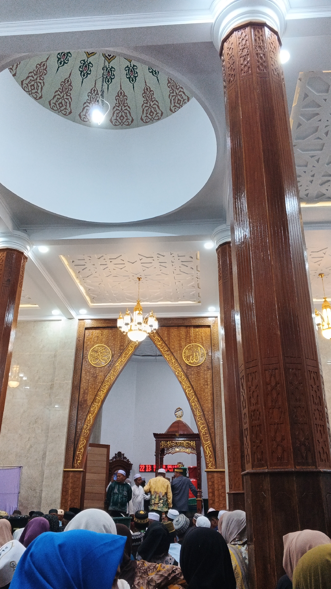 An indoor vertical shot captures a congregational prayer at a mosque in Pejala, Indonesia. A large, ornate wooden pillar dominates the right side of the frame, extending upwards towards a decorative ceiling. In the center, a grand archway made of intricately carved wood leads to the mihrab, the prayer niche. Within the archway, gold Arabic calligraphy adorns the wooden structure. Above the mihrab, a smaller wooden structure with a dome is visible. A chandelier with multiple lights hangs from the ceiling. To the left of the archway, a segment of the mosque's dome is visible, featuring decorative patterns. A digital display shows the time "22 13" and "A 06:49". In the foreground and midground, a congregation of people, mostly men wearing peci hats or skullcaps, are engaged in prayer. Some individuals are standing, while others are seated. The overall lighting is warm and ambient, illuminating the rich details of the mosque's interior.