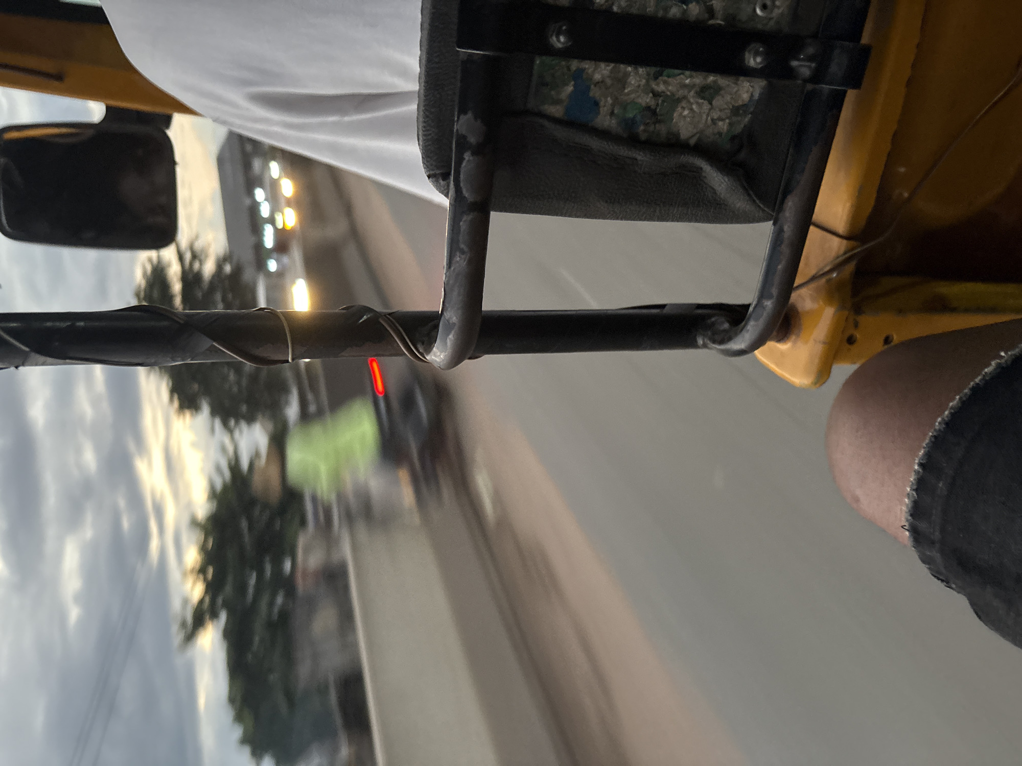 The image is taken from the interior of a yellow motorized tricycle, commonly known as a "keke" or "Marwa" in Nigeria, suggesting it is likely operating within the city of Onitsha, Nigeria. The perspective is from a passenger's viewpoint, looking forward and to the side.
In the foreground, a person's leg, clad in dark denim shorts, is visible on the left. The seat of the tricycle is also visible, upholstered in a dark, possibly faux leather material, with a section showing a colorful, patterned fabric underneath. A black metal frame, part of the tricycle's structure, runs across the frame.
The middle ground shows a road with blurred motion, indicating the tricycle is in transit. On the left side of the road, a concrete barrier is visible. Beyond the barrier, a person in a bright green vest is on a motorcycle, also blurred due to motion. Trees with sparse foliage line the background. Further in the distance, on the right side of the road, a building or structure is visible with several blurred lights, suggesting other vehicles or illuminated signs. The sky is overcast with streaks of yellow and white, indicative of either sunrise or sunset. The overall lighting suggests it is either early morning or late evening.