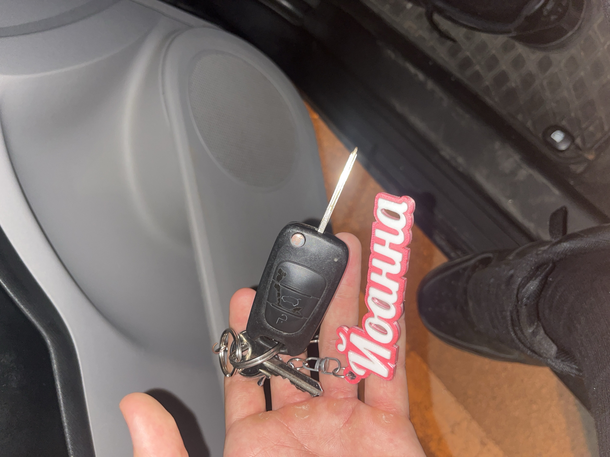 A first-person perspective photograph shows a hand holding a car key and a personalized keychain. The key is black and has a metal blade exposed. The keychain is pink and white and features the name "Йоанна" (Ioanna) in Cyrillic script. The background includes a light gray car door panel with a speaker grille and the bottom of a black shoe. Based on the Cyrillic script on the keychain, the location may be within a Cyrillic-script-using country. There is no way to determine the location specifically as Blagoevgrad, Bulgaria from the image.
