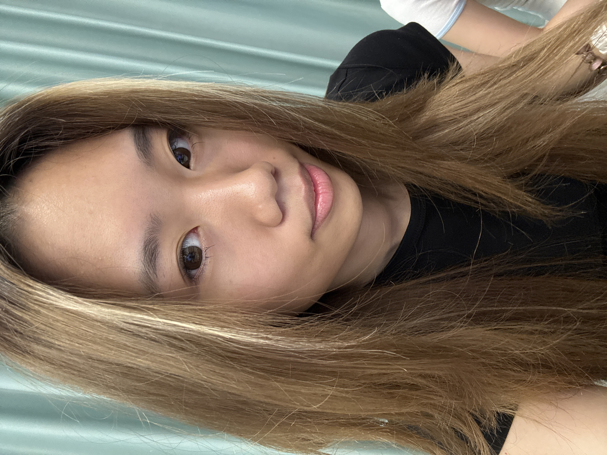 A portrait of a young woman with long, light brown hair is centered in the frame. She is wearing a black top and looking directly at the camera with a neutral expression. Her skin tone is fair, and her lips appear slightly pink. The background is a blurry, light green color, suggesting an interior setting, possibly a room with curtains or a wall. A glimpse of a white garment can be seen towards the top-right corner of the frame.
This image does not contain sufficient information to identify a precise location within Nan'an District, China.