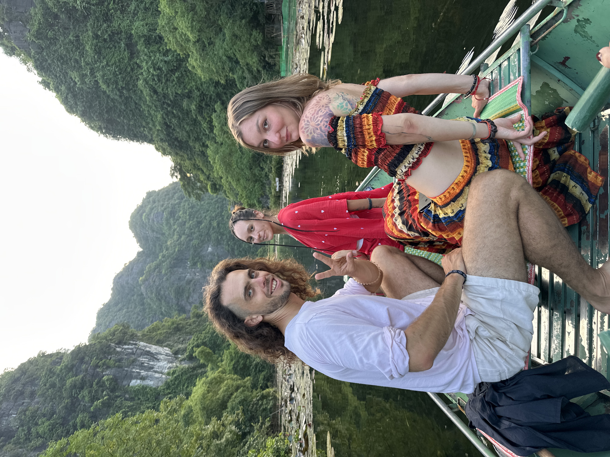 Three individuals are seated in a boat on a body of water surrounded by karst mountains in Nam Hoa Lư Ward, Vietnam. The person on the left is a male with long, curly brown hair, wearing a white t-shirt and light-colored shorts. He is smiling and holding up two fingers in a peace sign. The person in the middle is a female with dark hair braided down her back, wearing a red robe. The person on the right is a female with blonde hair, wearing a colorful, off-the-shoulder top and skirt. She has tattoos on her arm and is looking over her shoulder towards the camera. The boat appears to be a traditional wooden vessel, and the water is covered with lily pads. The mountains in the background are densely vegetated.
